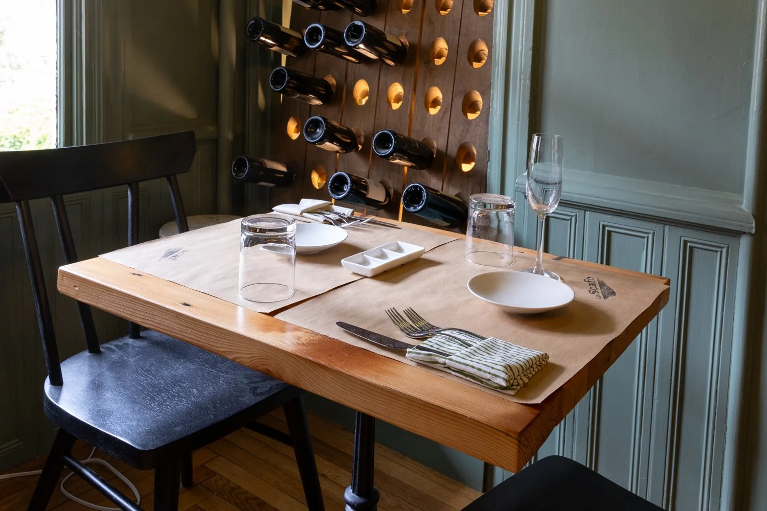 A dining table set with glassware, bowls, and utensils in a room with green paneled walls and a wine rack with bottles on the wall.