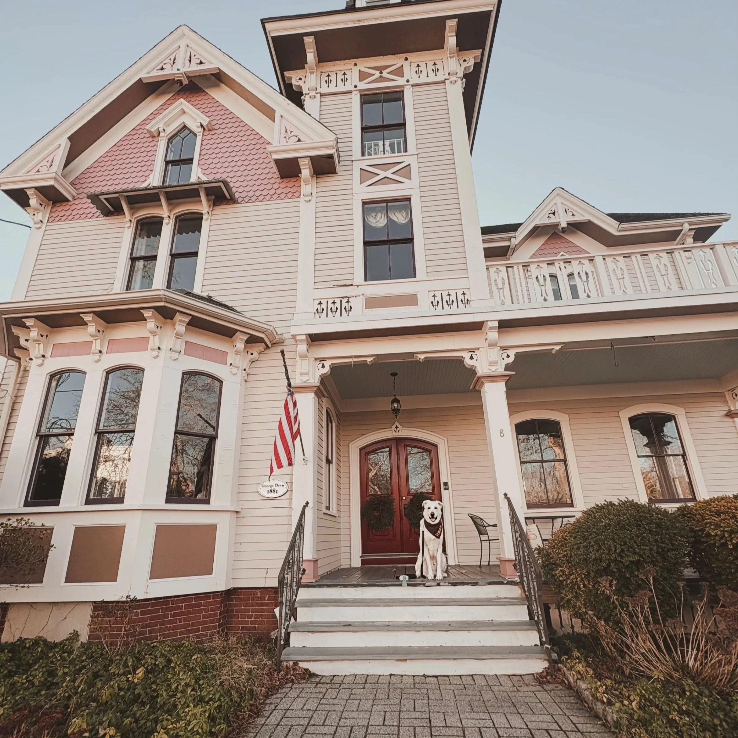 Front view of an ornate Victorian-style house with a porch, multiple windows, decorative trim, and a flag hanging by the door. A dog is sitting on the steps in front of the house.
