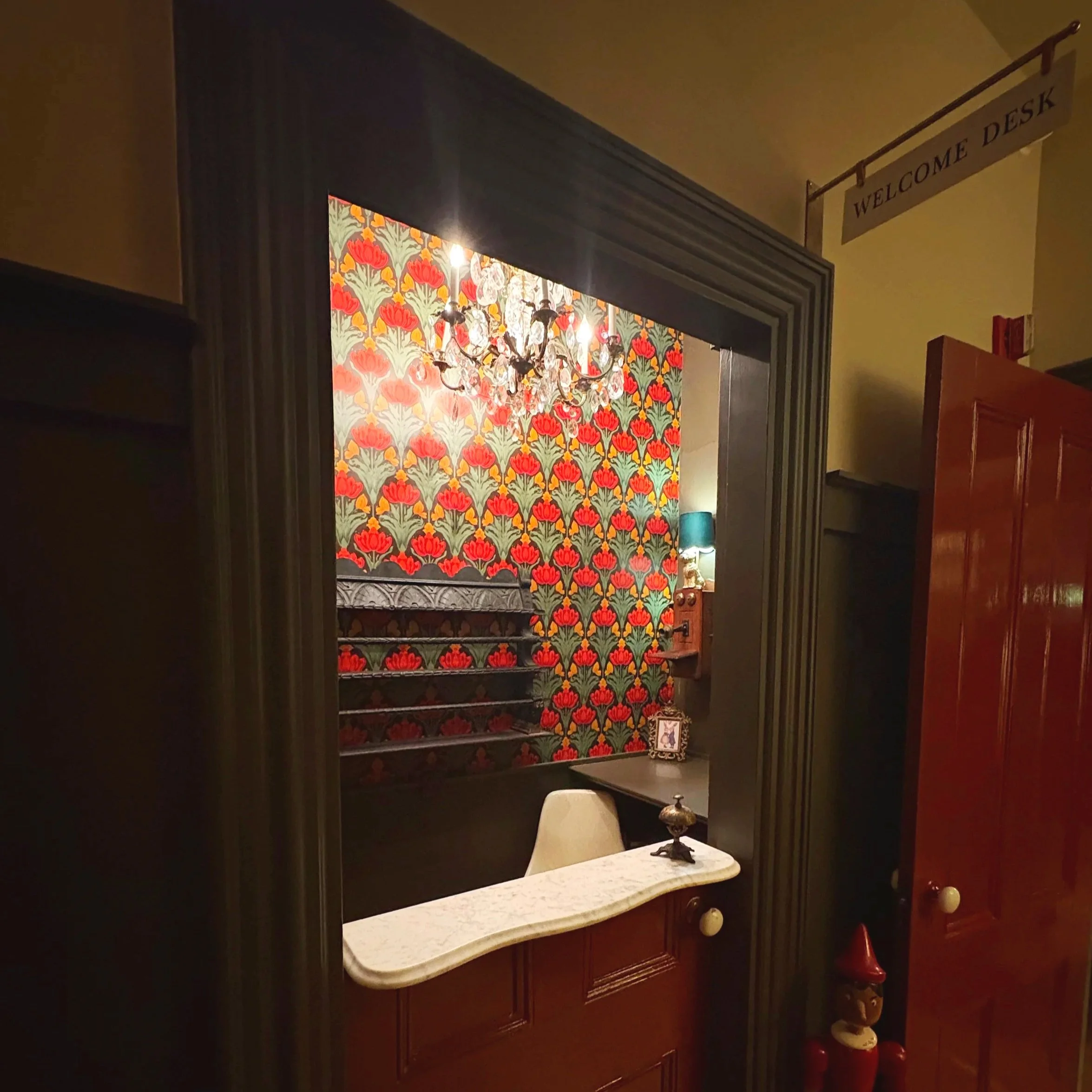 Interior of a room with a floral wallpaper, chandelier, and a white counter with a decorative lamp. A 