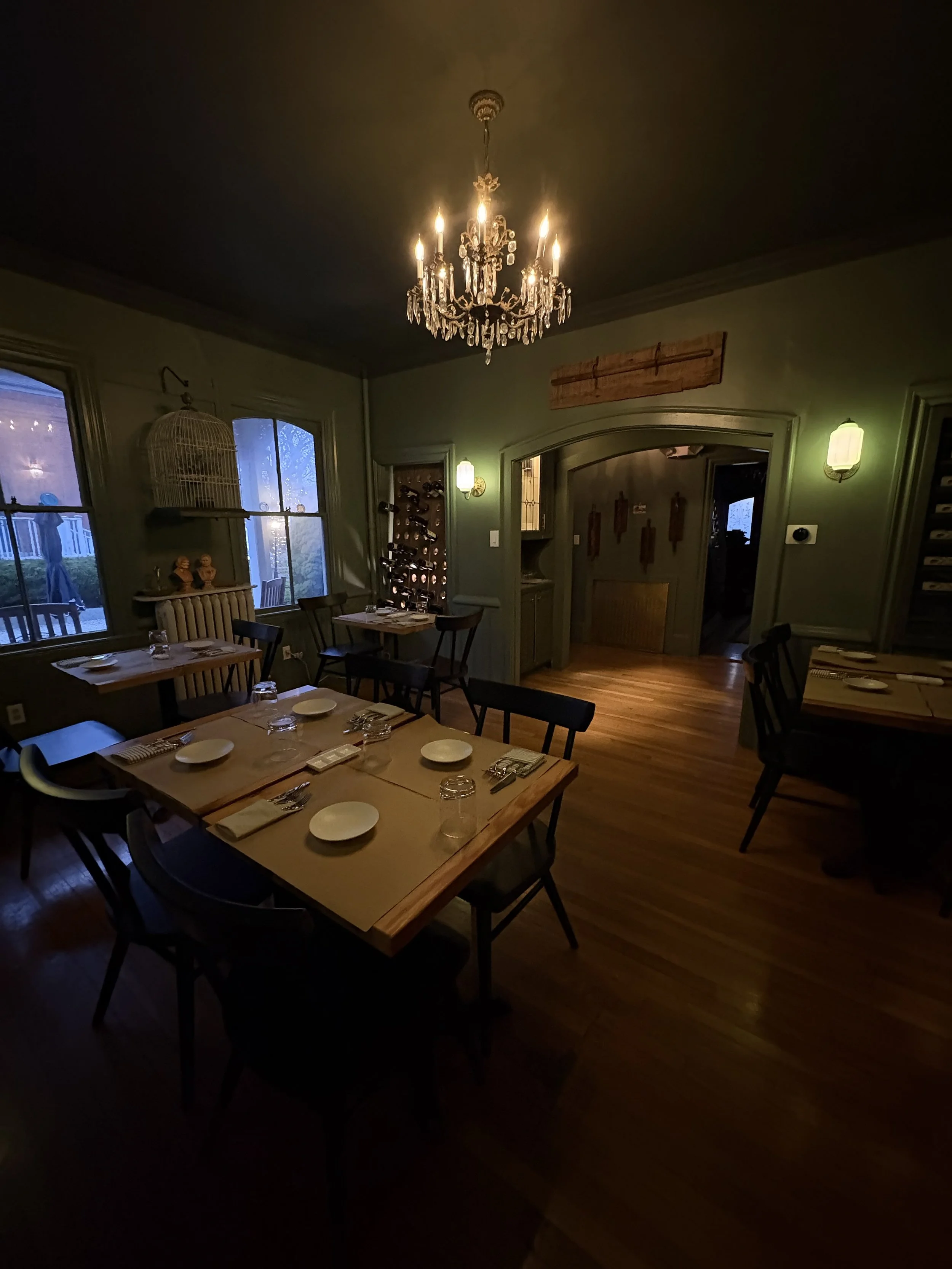 A cozy dining room with a chandelier hanging from the ceiling, wooden tables set with plates and glasses, and windows showing an outdoor scene.