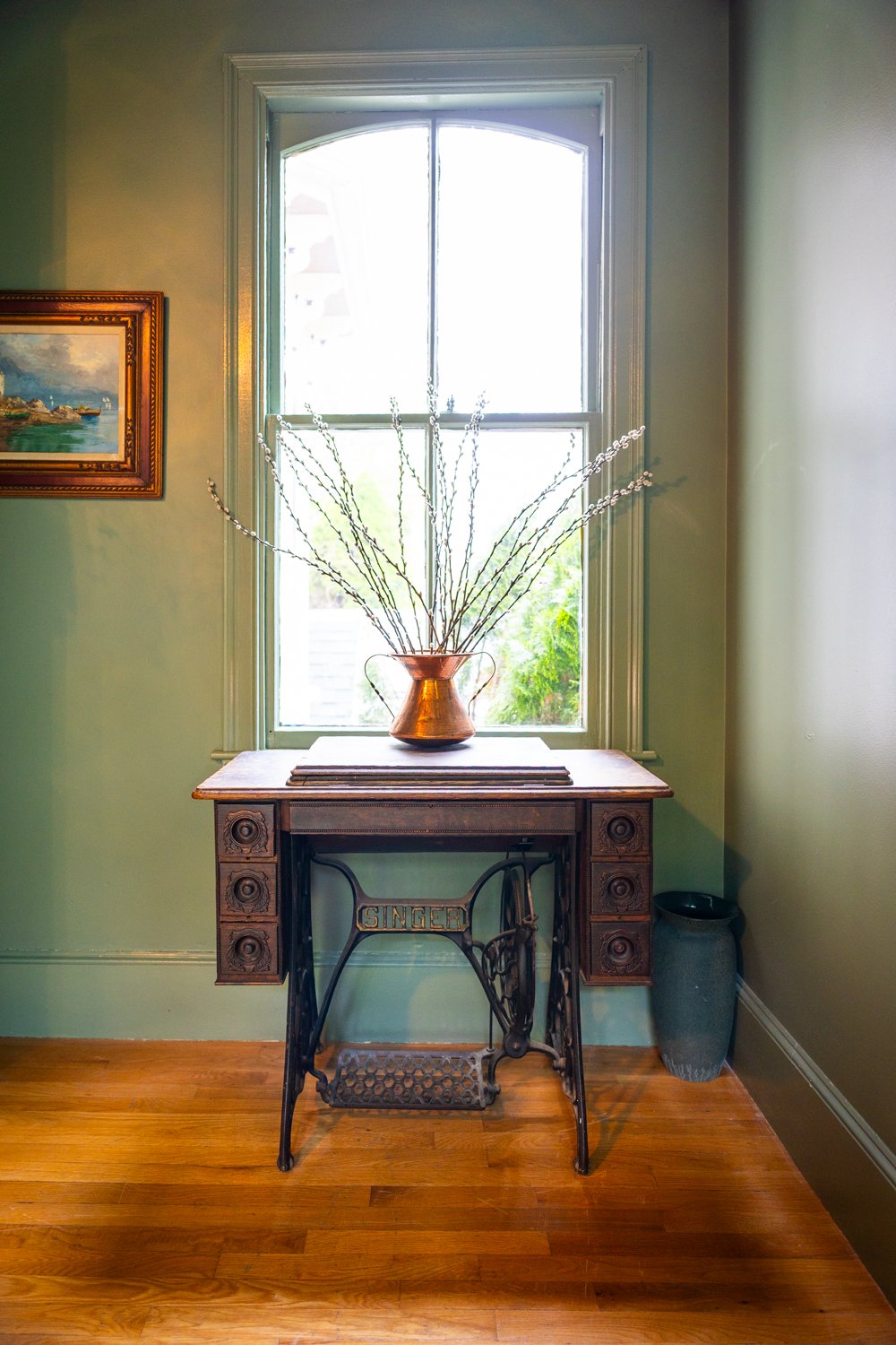 A vintage Singer sewing machine repurposed as a table with a wooden top, holding a copper vase with long, thin branches in front of a window with white trim, in a room with wooden flooring and light green walls. A framed painting hangs on the left wa