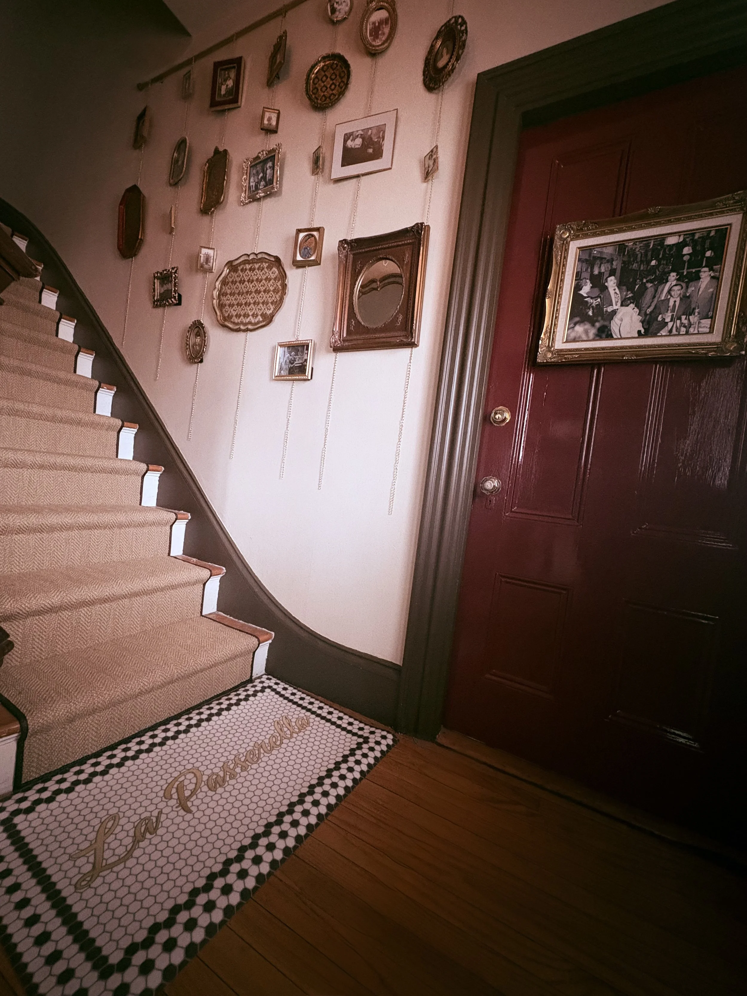 A staircase with a patterned carpet runner next to a wall decorated with various framed pictures and decorative plates. There is a large framed photograph on a red door to the right and wood flooring in the foreground.