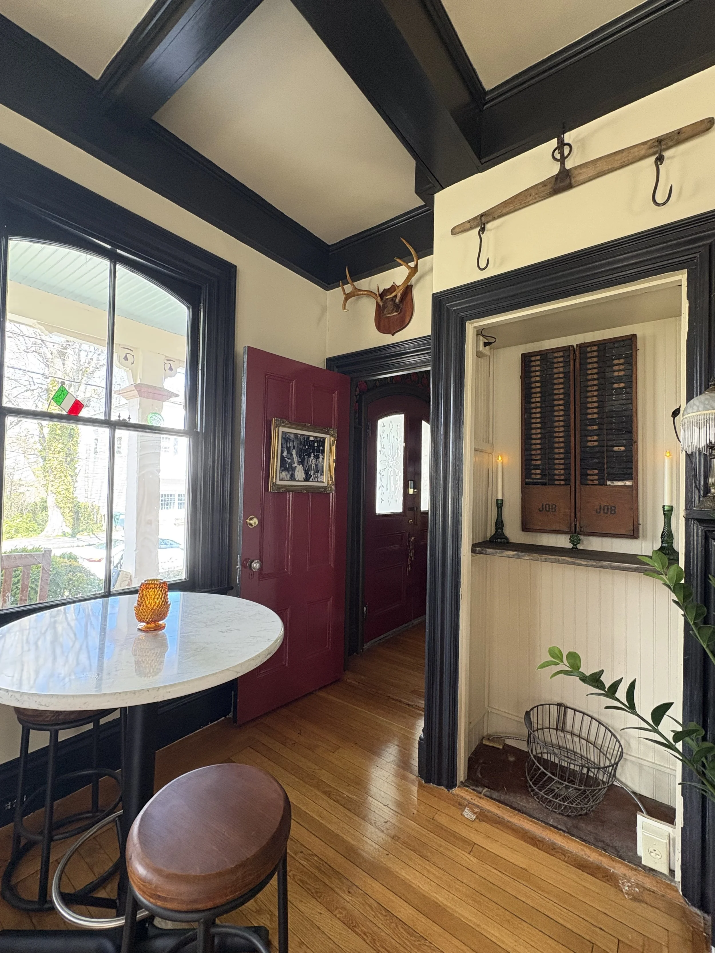 Interior of a room with a window, a red door, a table with an orange candle, wooden floors, and decorative items including a mounted deer head and hooks on a wooden rod.