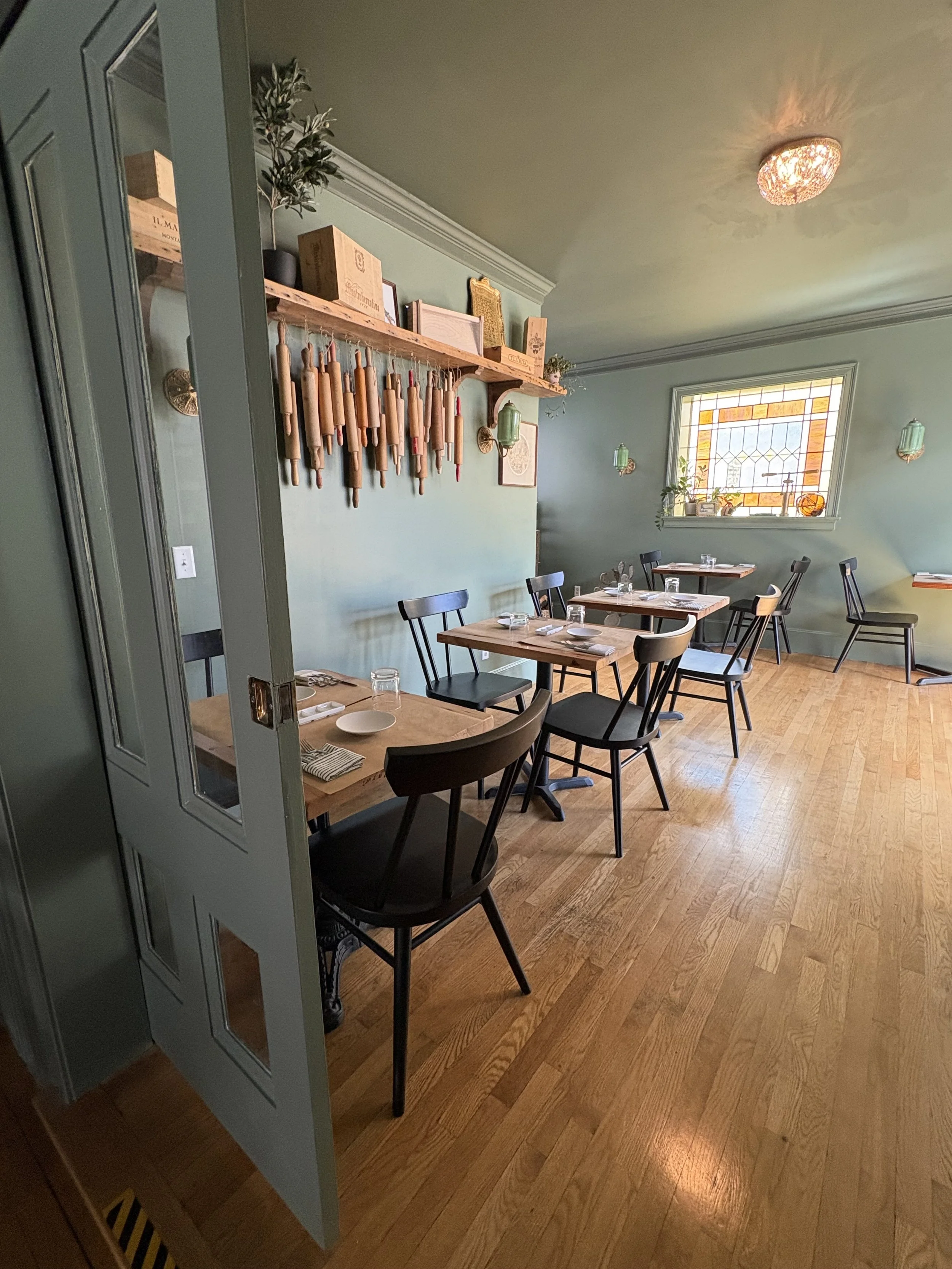 A cozy restaurant interior with light green walls, a wooden floor, tables set with glasses and plates, black chairs, a large window with stained glass, and a shelf with decorative items and hanging kitchen utensils.