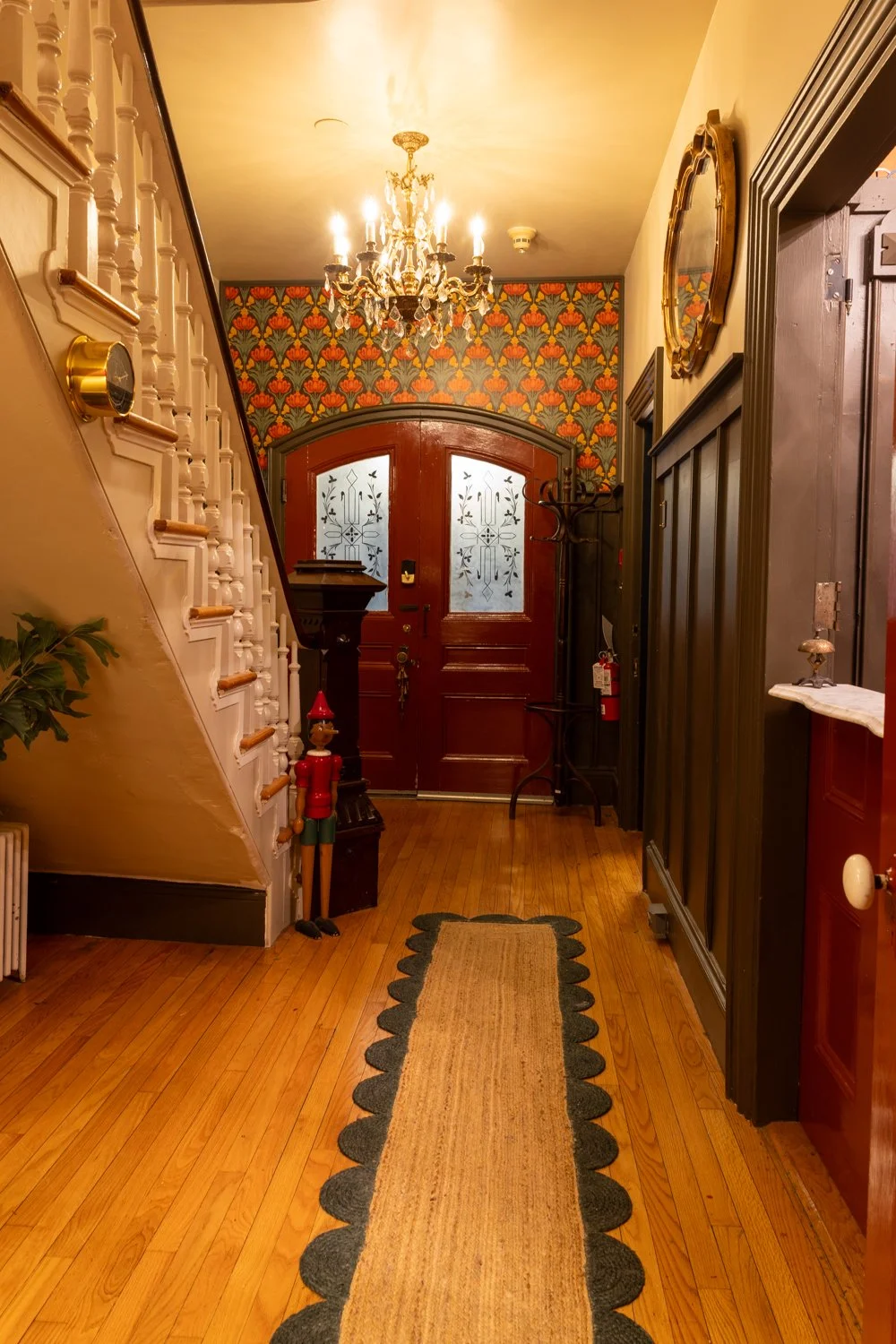 A hallway with a wooden floor and a runner rug, a staircase on the left, a chandelier hanging from the ceiling, a coat rack, decorative wallpaper on the wall, and a double front door with frosted glass panels.