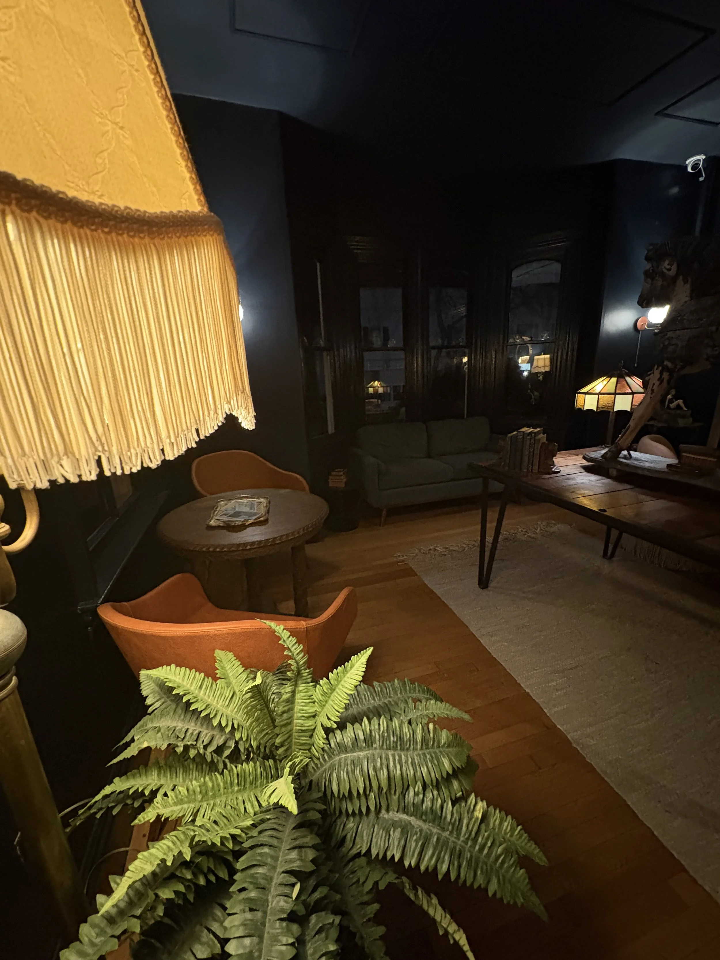 Living room with a large fern plant in the foreground, a vintage orange chair, a wooden table with magazines, green cushioned sofa, dark wood-paneled walls, large windows with black trim, and ambient lighting from a stained glass table lamp and wall 