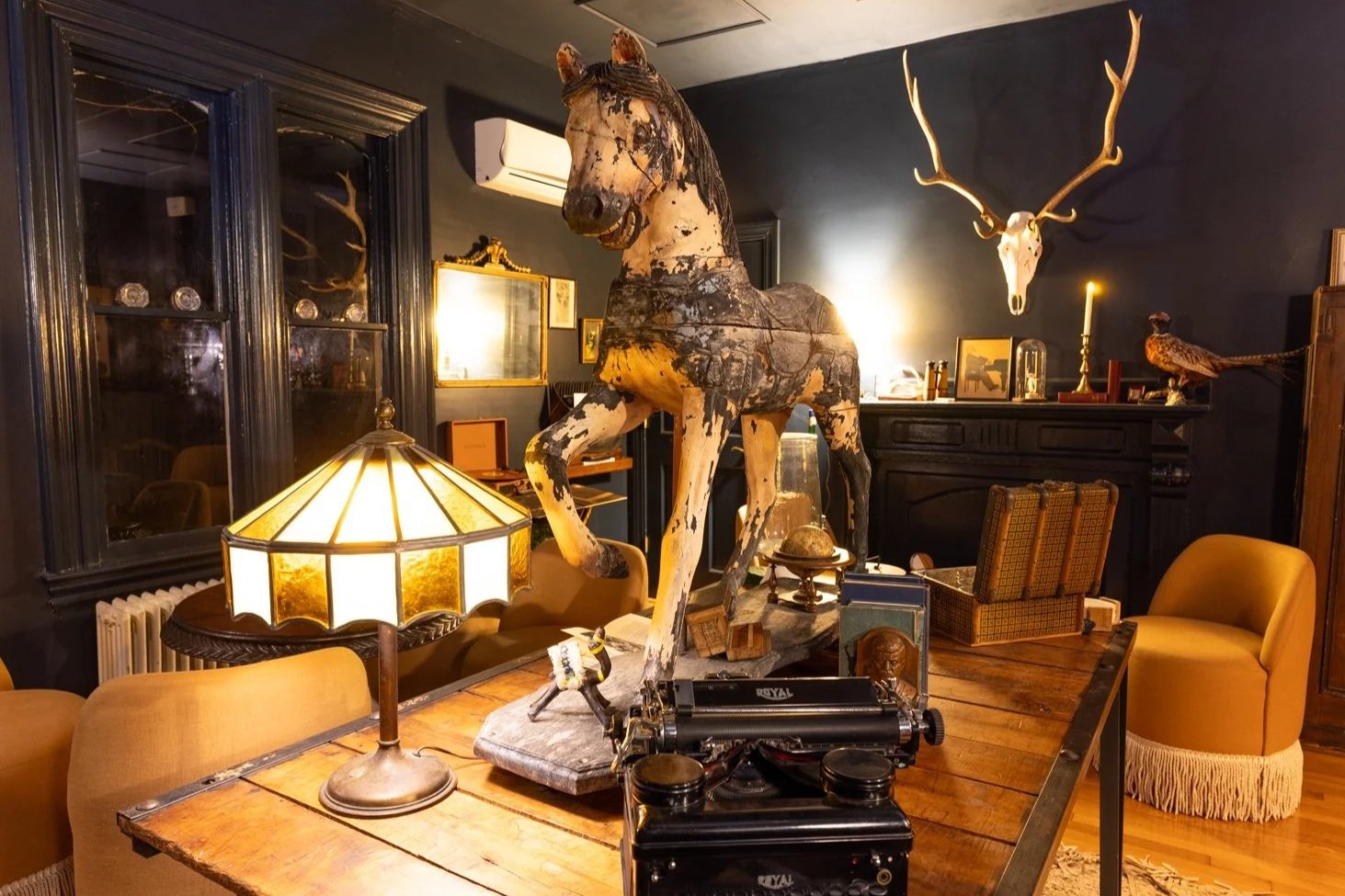 Decorative rustic room with vintage and taxidermy items including a horse head and a reindeer skull with antlers on dark blue walls, antique furniture, and a table lamp with stained glass shade.