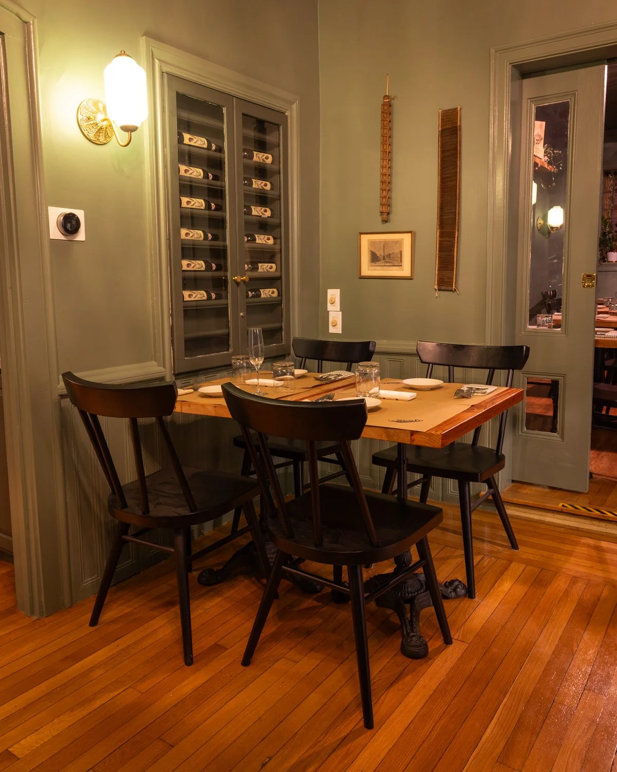 A wooden table set for four with plates, napkins, glasses, and silverware in a cozy dining room with green walls, a wine cabinet, and decorative wall hangings.