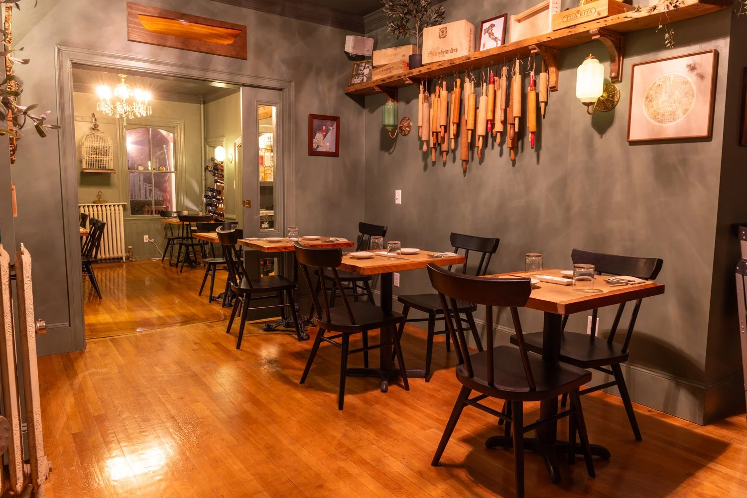 Cozy restaurant interior with wooden floors, green walls, and modern furniture. Small tables are set with glasses, plates, and utensils. The wall decoration includes framed art, hanging wooden kitchen tools, and vintage-style lighting fixtures. A doo
