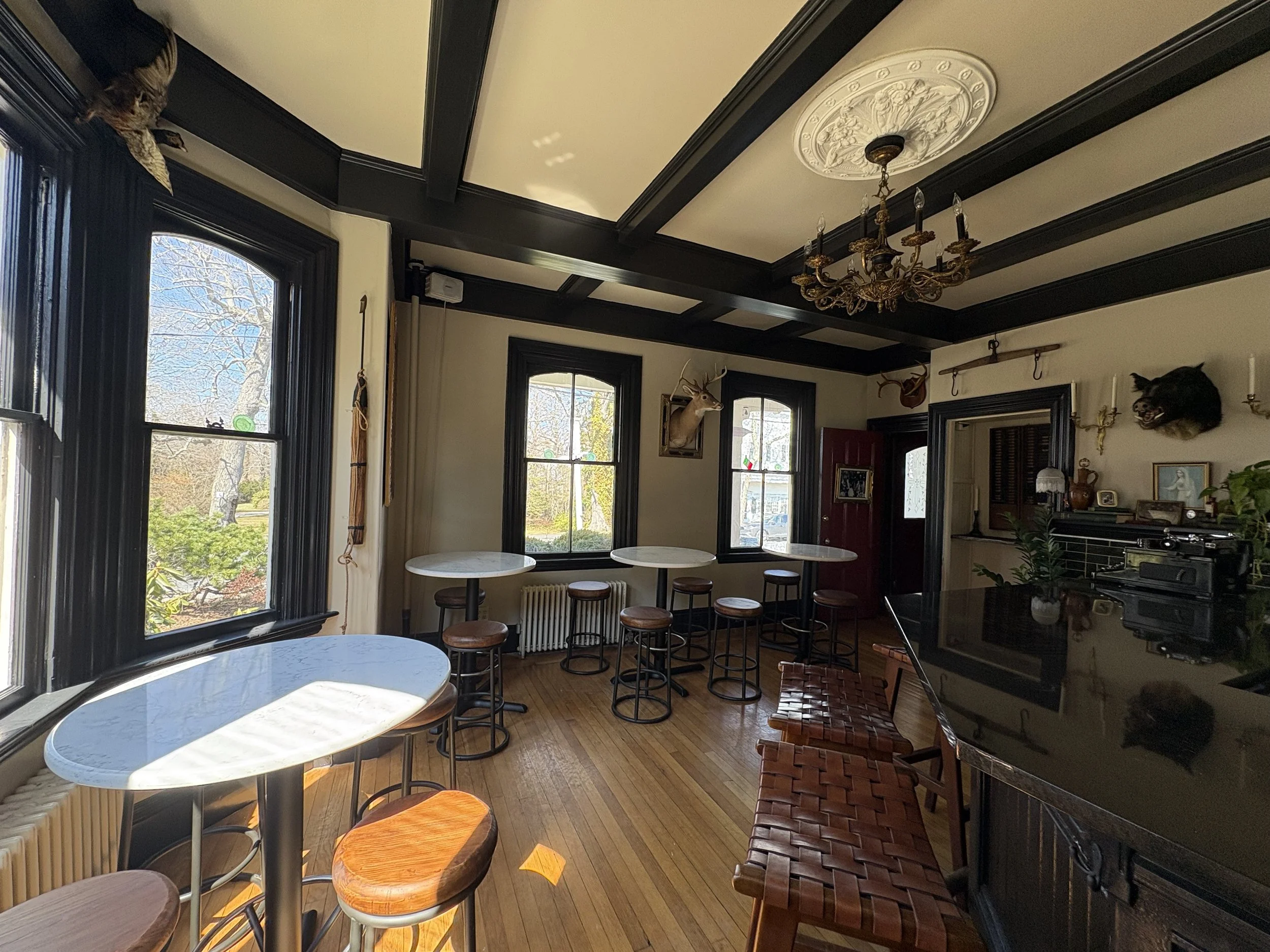 A cozy dining area with large windows, round marble tables, tall bar stools, and wall-mounted animal heads, including a deer and a pig, in a room with black trim and hardwood floors.