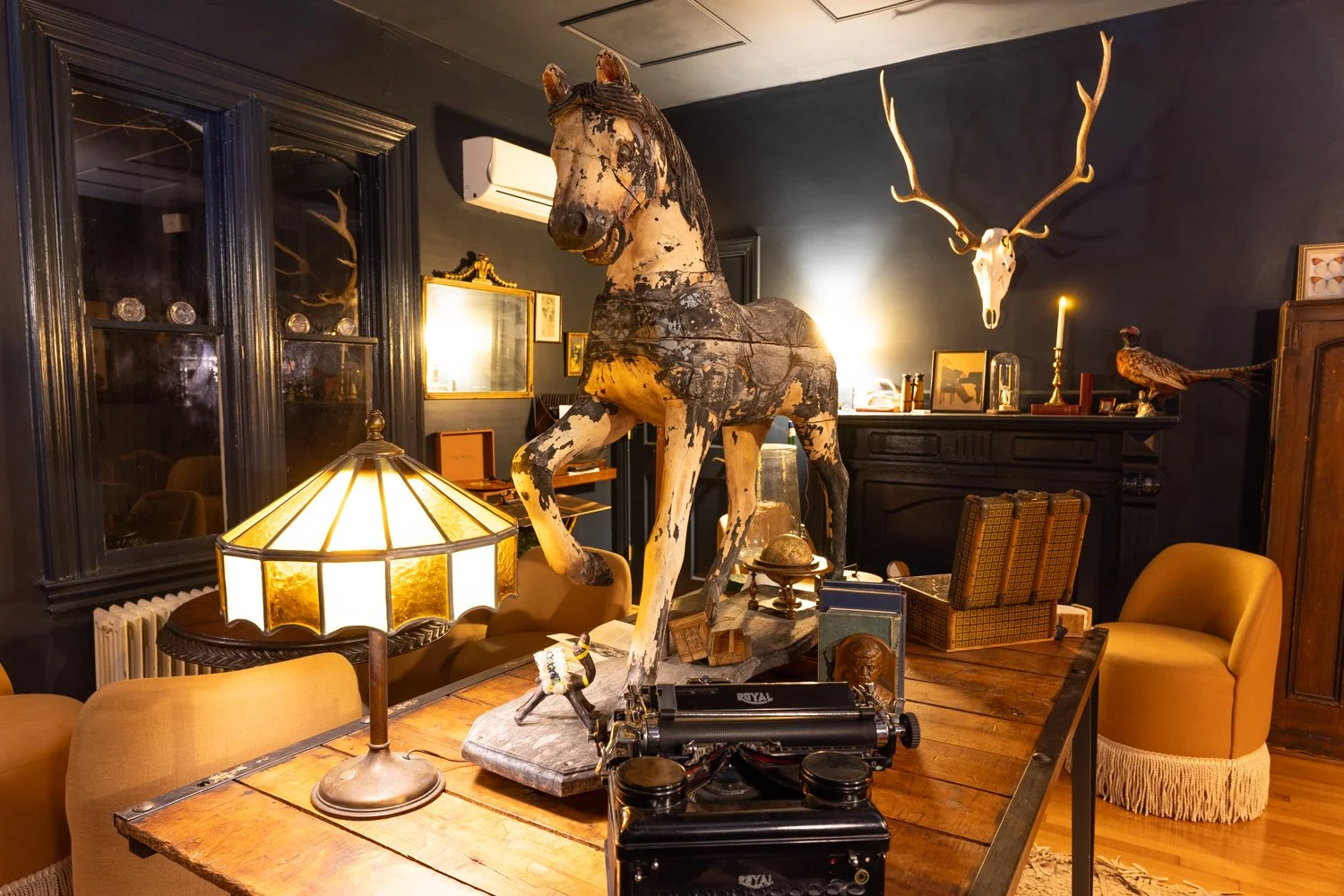 Decorative room with antique furniture, mounted animal and skulls, and vintage items including a horse sculpture, an elk skull, and a typewriter, illuminated by a vintage table lamp.