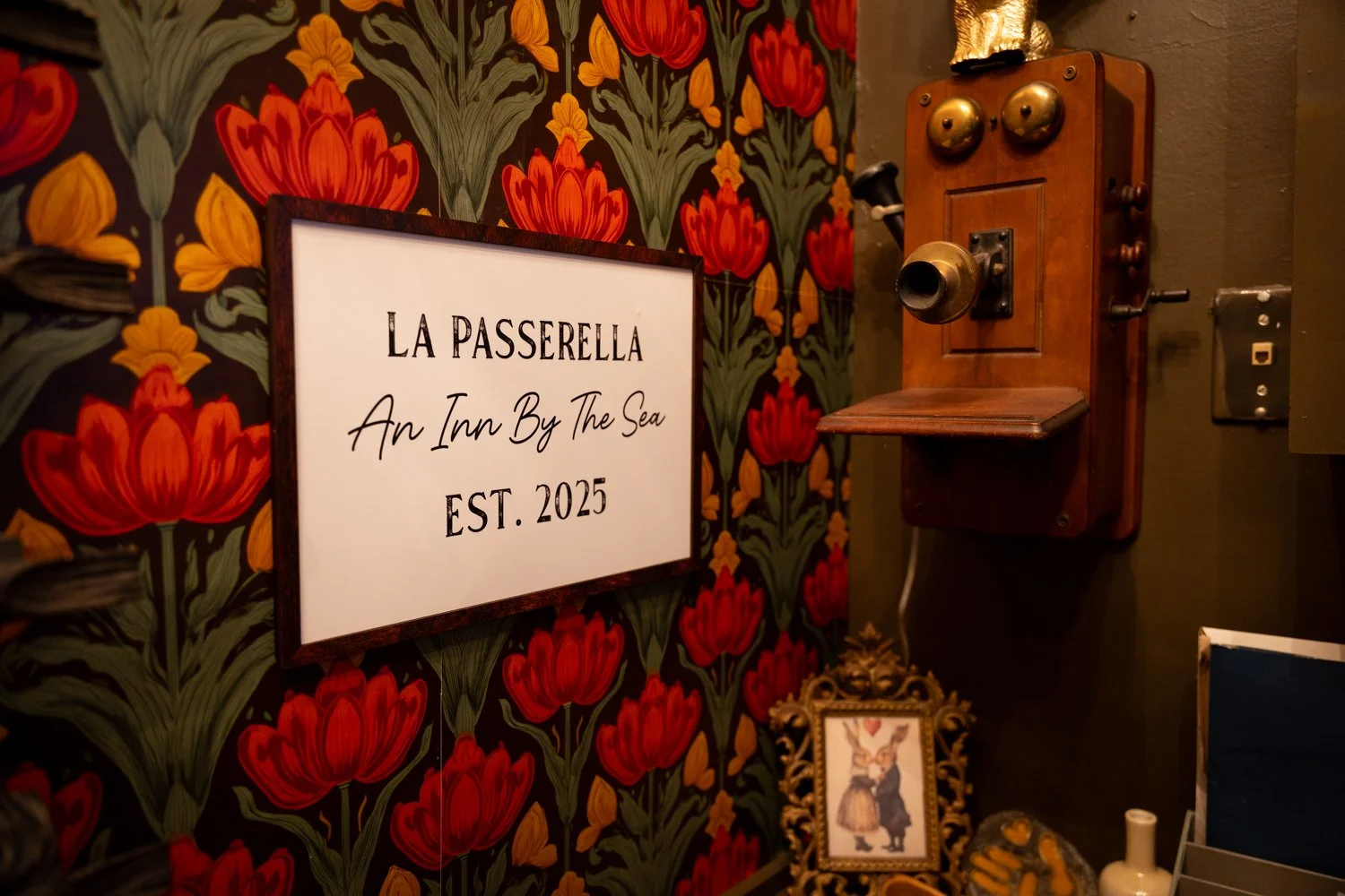 Decorative interior wall with a floral patterned wallpaper featuring red and orange flowers, a framed sign that reads 'LA PASSERELLA, An Inn By The Sea, EST. 2025,' a vintage wooden wall telephone, and a small framed picture of a rabbit dressed in cl