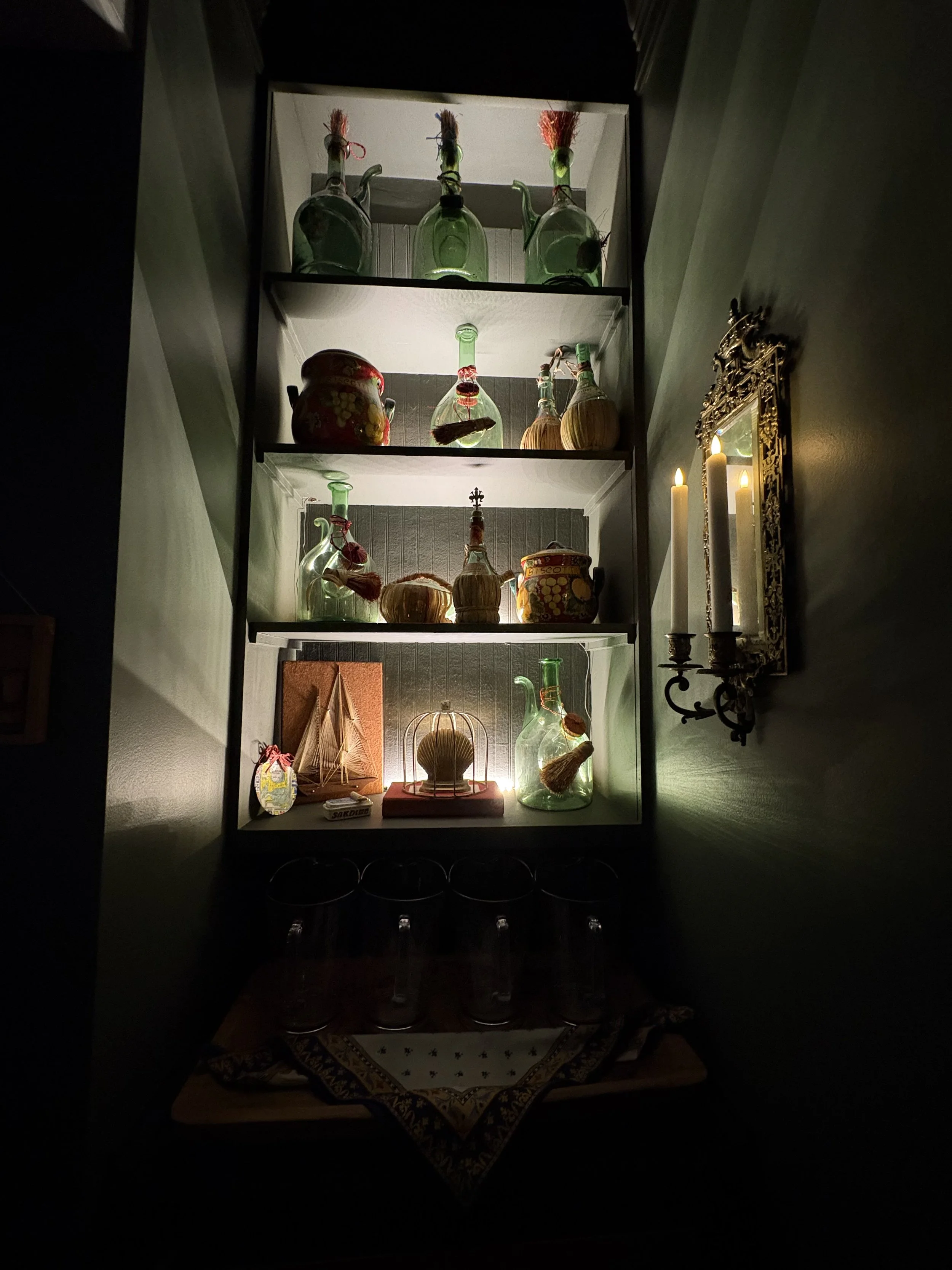 A dark cabinet with four shelves, each illuminated from within, displaying various glass bottles, jars, and decorative items. To the right of the cabinet is a mirror with an ornate gold frame and three lit candles.