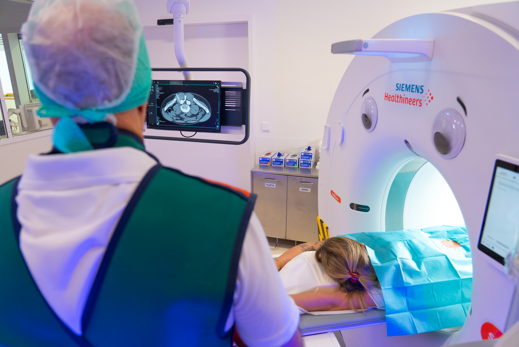 Arzt steht vor einem CT-Scanner, während ein Patient auf dem Bett liegt, bereit für die Untersuchung, in einem medizinischen Raum.