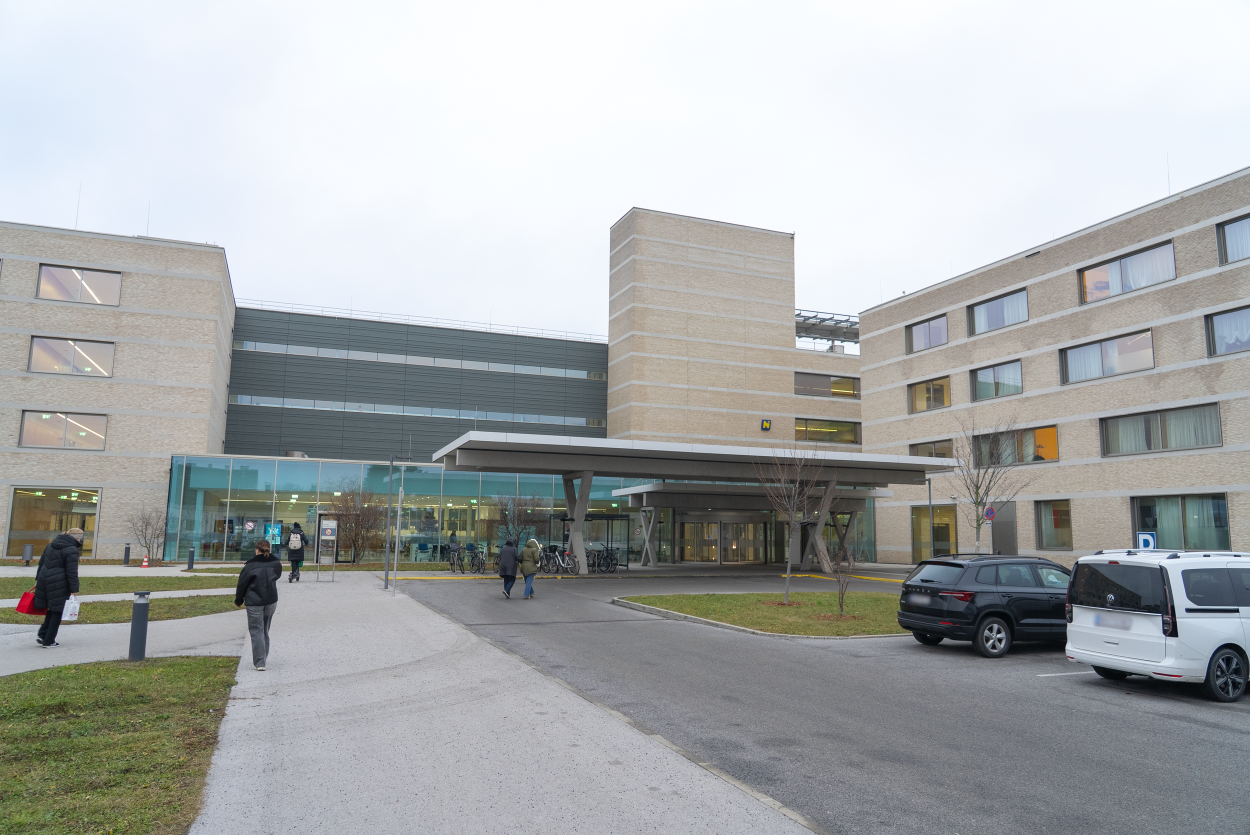 Ein modernes Krankenhausgebäude mit mehreren Etagen und großen Fenstern, Menschen gehen auf dem Gehweg vor dem Eingang, es gibt Parkplätze und Fahrradständer vor dem Gebäude.