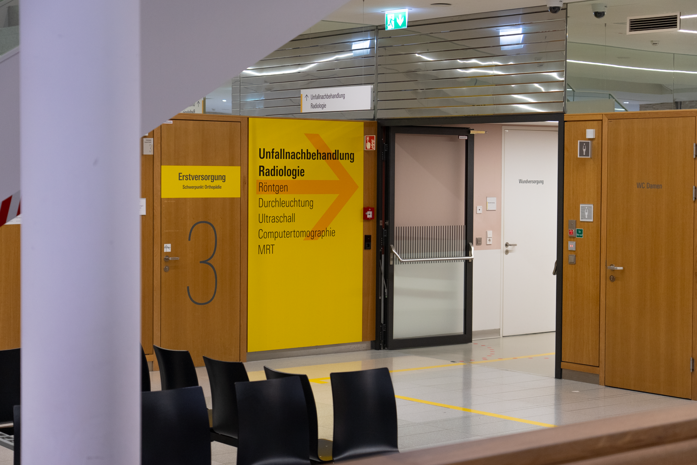 Bereich in einem Krankenhaus mit Schildern für Unfallnachbehandlung Radiologie, Erste Versorgung und Wartebereich mit schwarzen Stühlen.