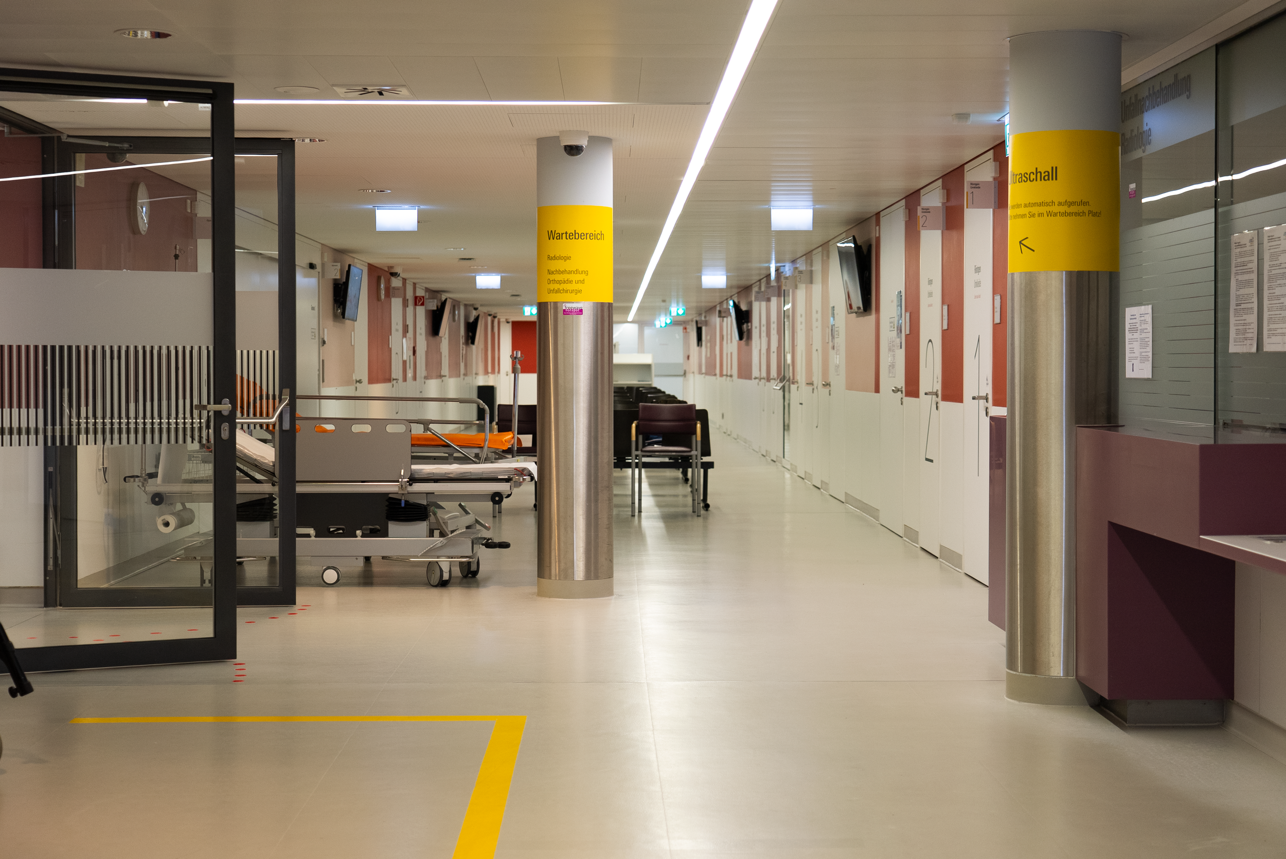 Leerer Krankenhausflur mit Rollbett und Stühlen, gelben Hinweisschildern und mehreren Monitoren an der Wand.