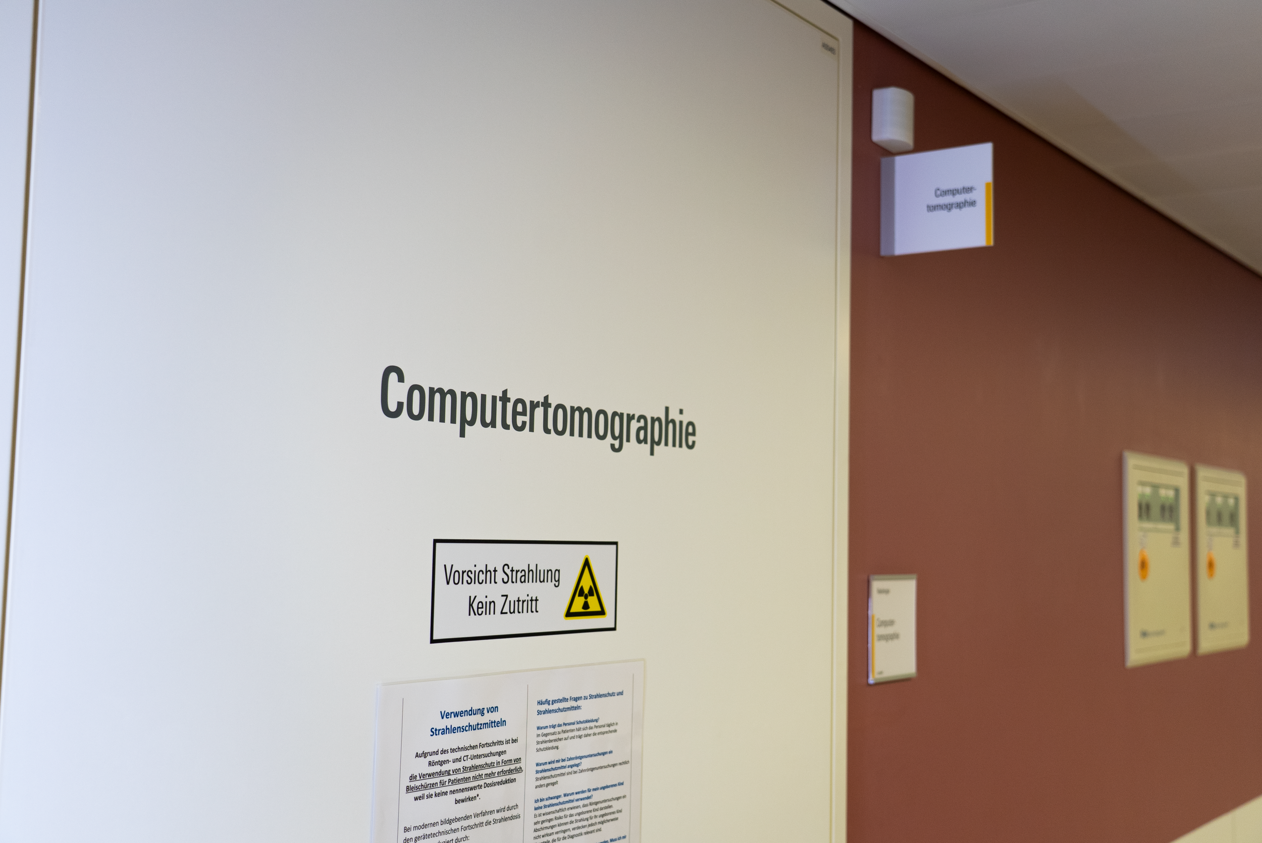 Hinweisschild an einer Wand mit der Aufschrift 'Computertomographie' und einem Warnsymbol. Zusätzliche Beschilderung und Informationstafeln sind sichtbar, in einem medizinischen oder klinischen Umfeld.