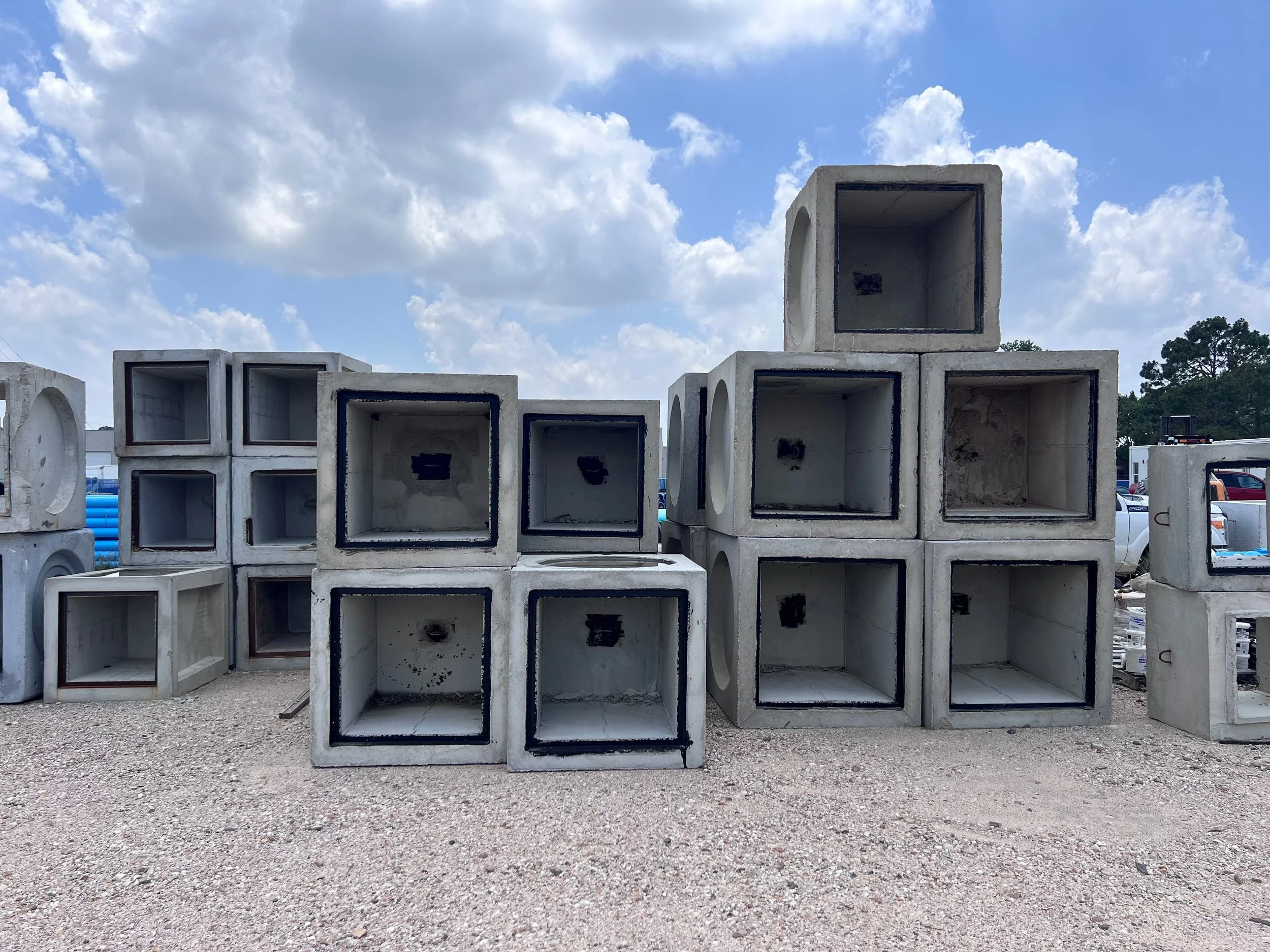 Stacked concrete pipes with black interior lining placed outdoors on a gravel surface under a partly cloudy sky.
