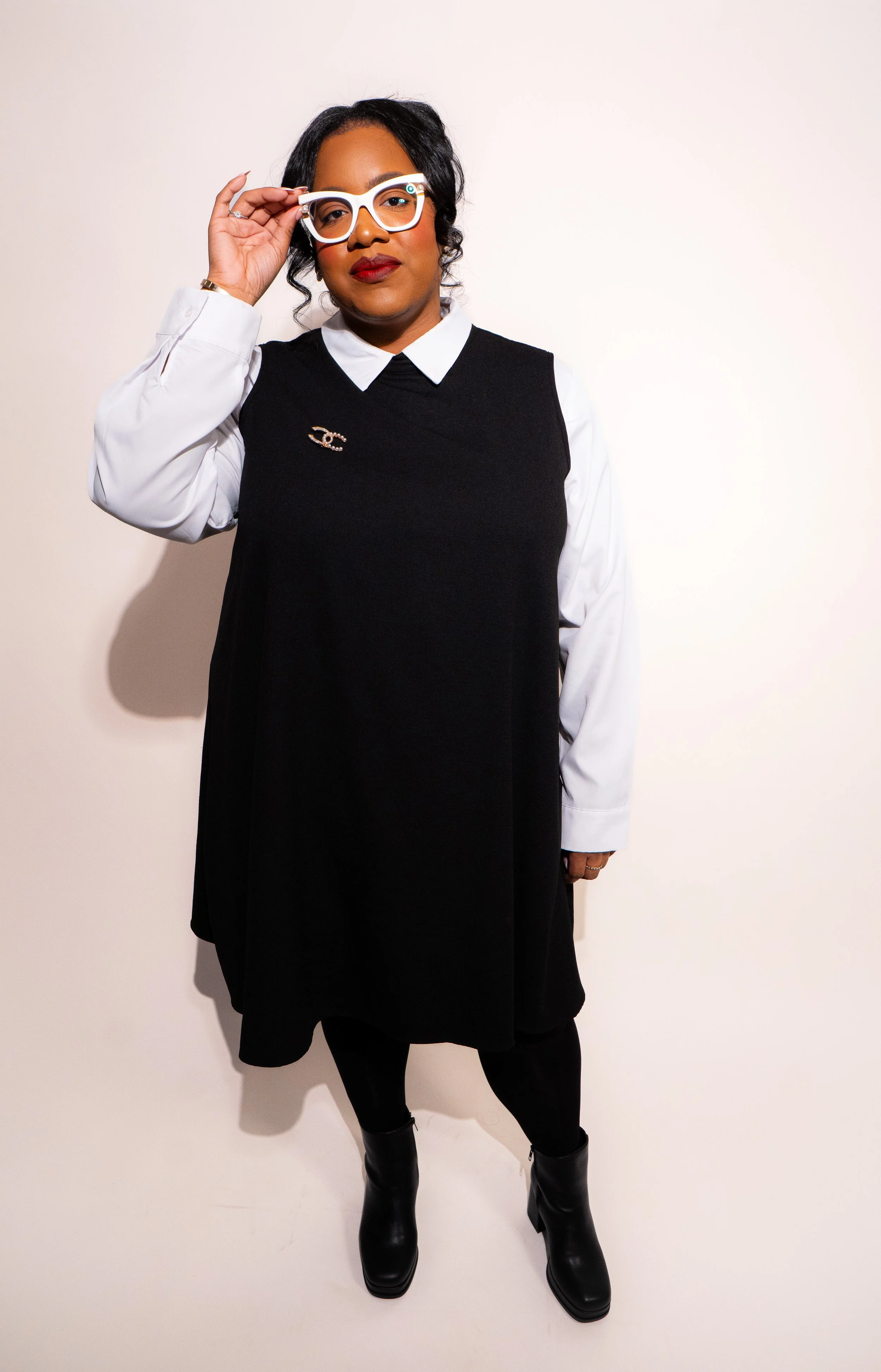 A woman with dark hair, wearing white glasses, a white collared shirt under a black sleeveless dress, black boots, and a Chanel brooch, standing against a plain off-white background.