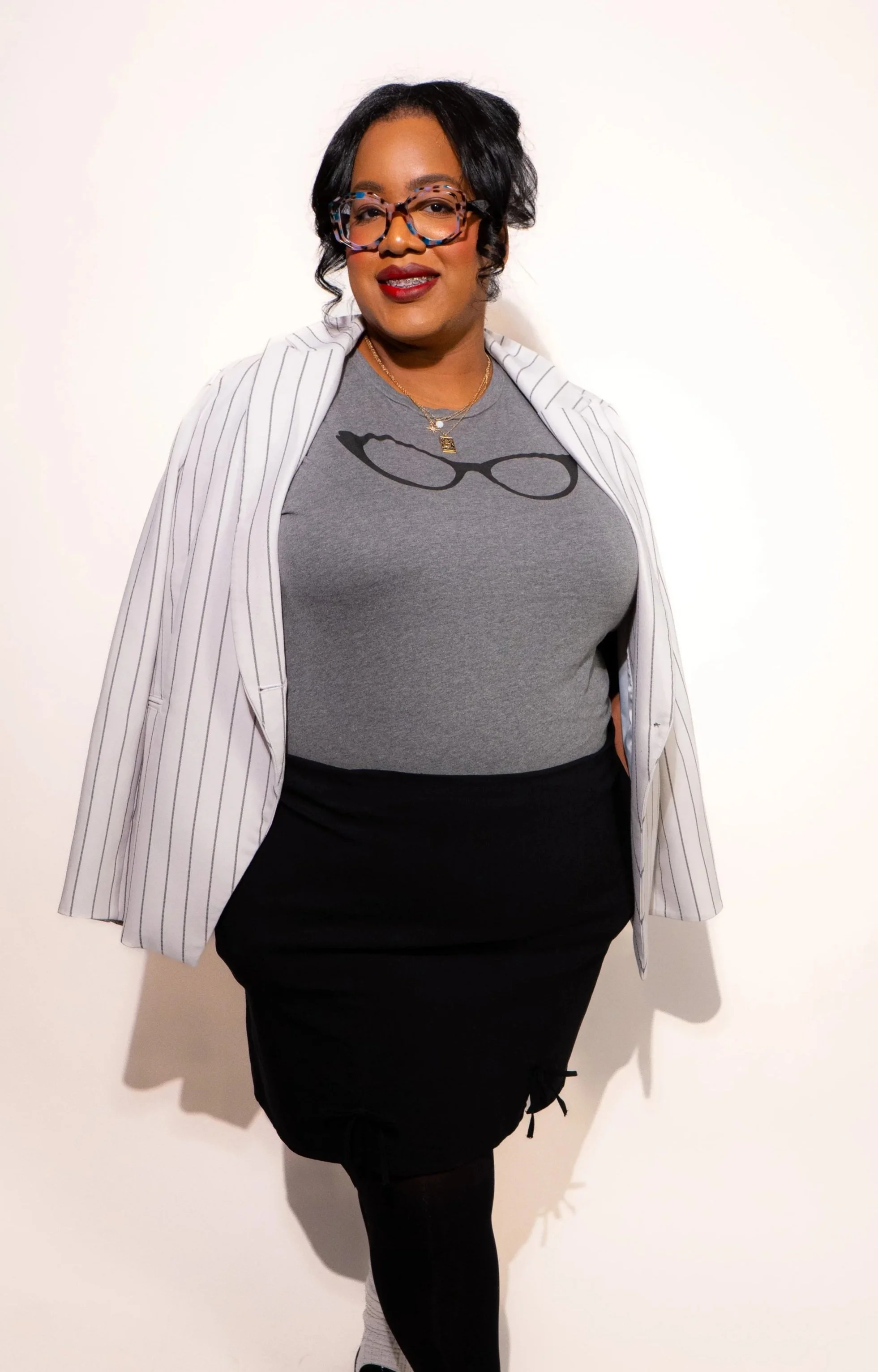 A woman with black hair wearing glasses, a gray graphic t-shirt, a white pinstripe blazer, and a black skirt, standing against a plain white background.