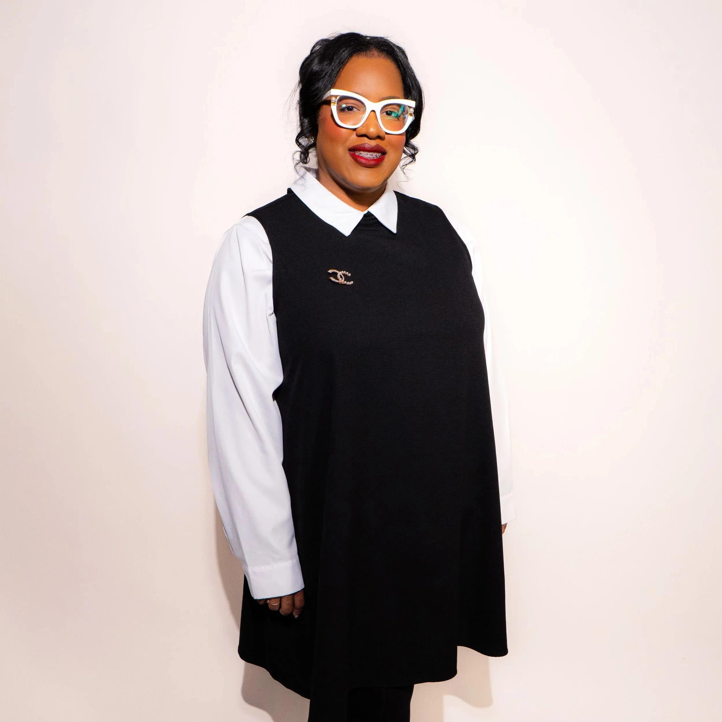 A woman with glasses standing against a plain white wall, wearing a black dress over a white shirt with a brooch.