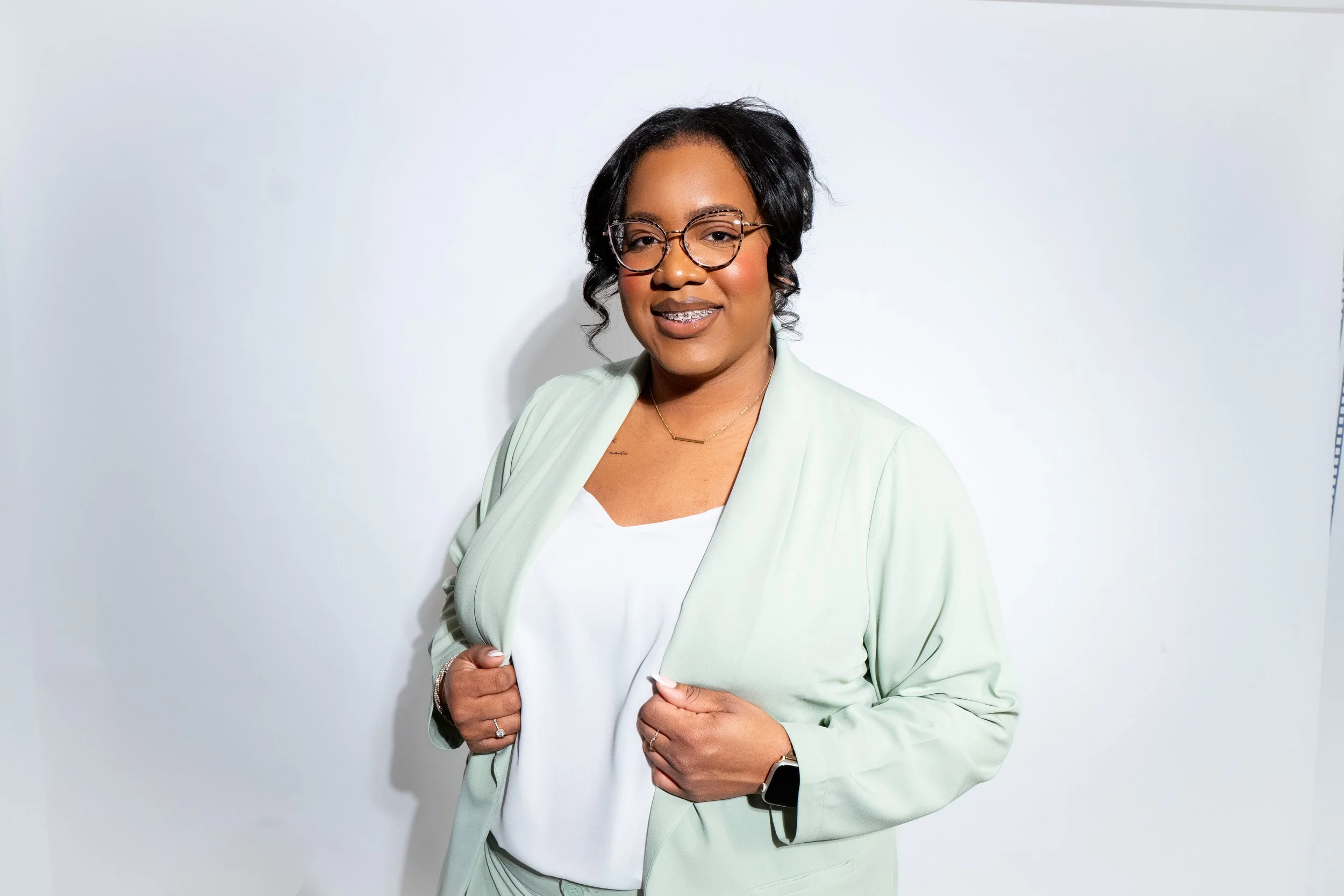 A woman with glasses, short black hair, and a warm smile, wearing a light green blazer over a white top, standing against a plain white background.