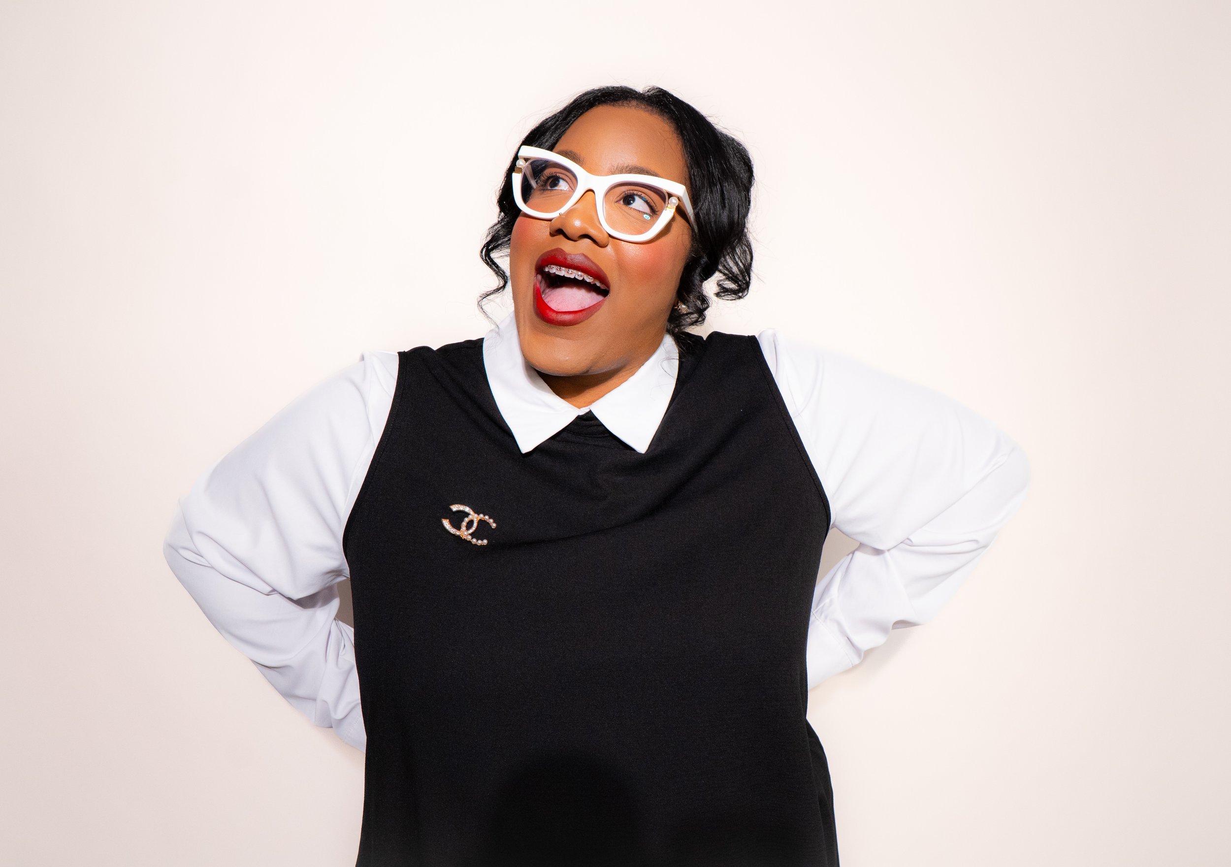 A woman with black hair, red lipstick, white glasses, wearing a black and white shirt with a Chanel brooch, posing with her hands on her hips and smiling.