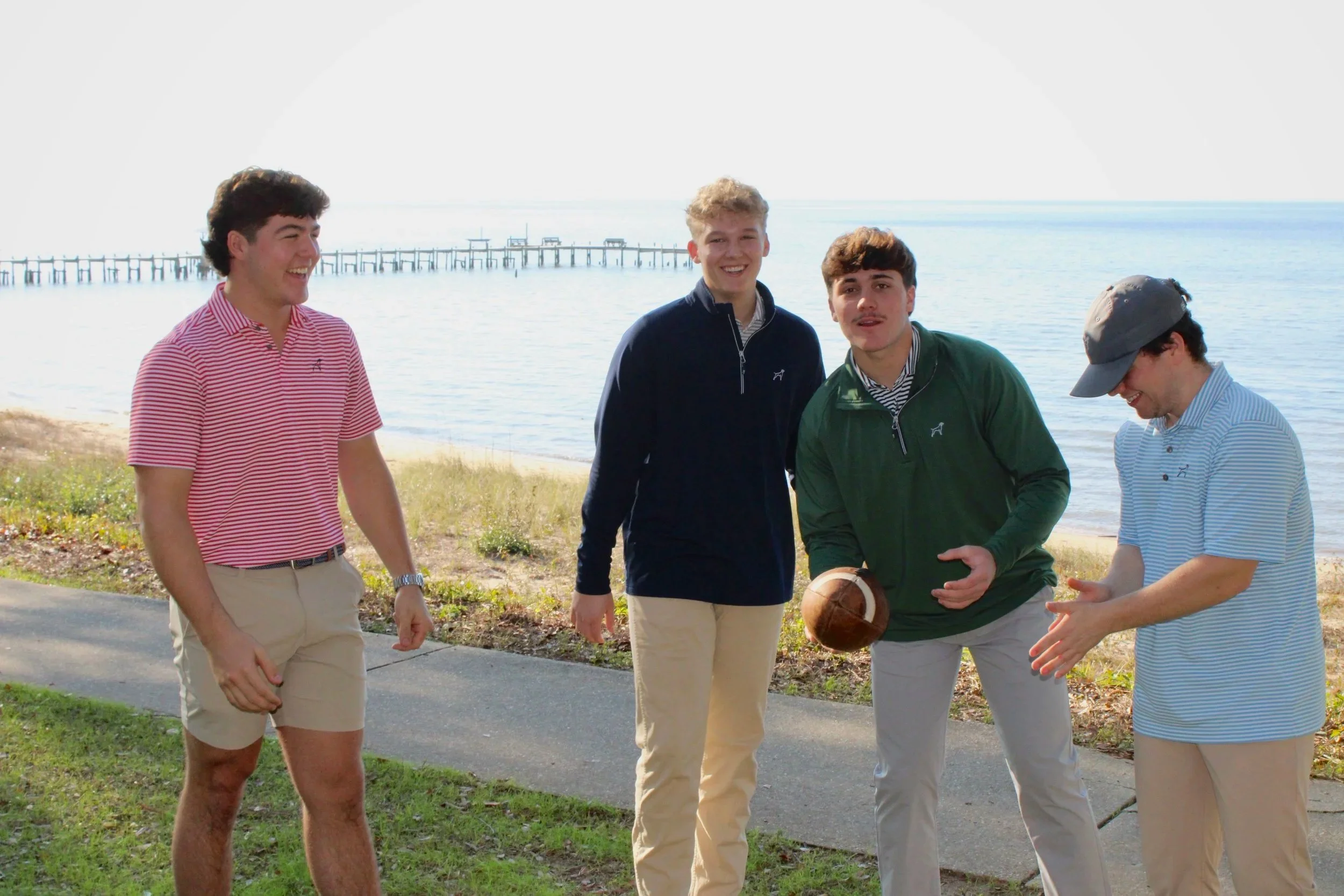 Four young men standing on a grassy area near a paved path by the water, smiling and talking. One is holding a football. In the background, there is a body of water with a pier extending into it.