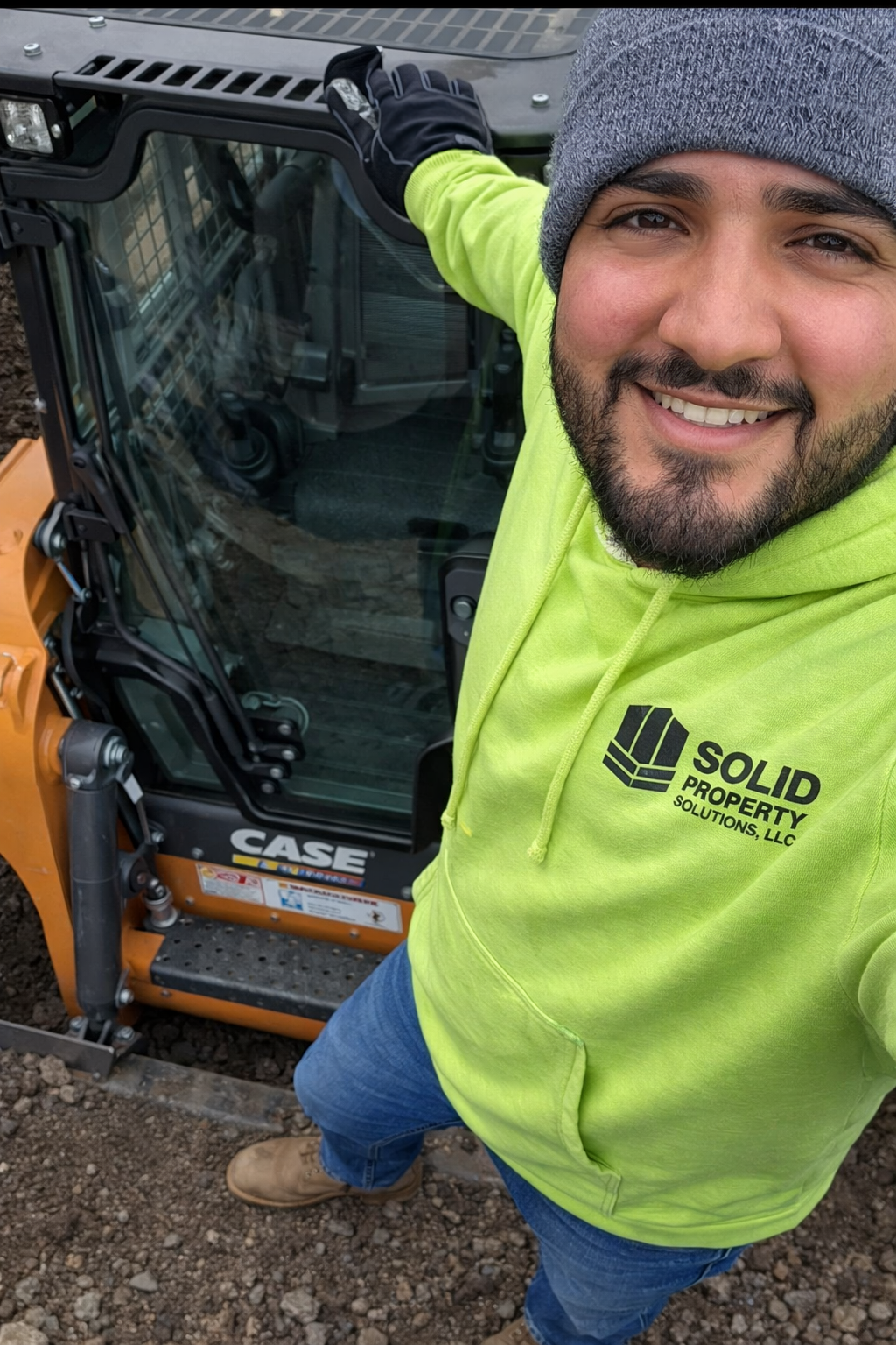 A man smiling while taking a selfie next to construction equipment, wearing a bright yellow hoodie with 'Solid Property Solutions LLC' logo, a gray beanie, and jeans.