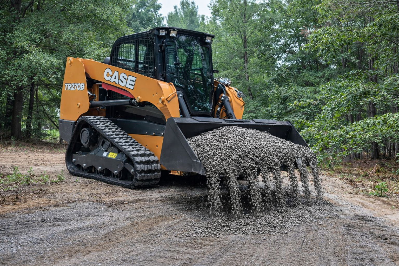 A yellow and black CASE compact track loader with a front bucket filled with small rocks, operating on a dirt path in a wooded area in Polk County.