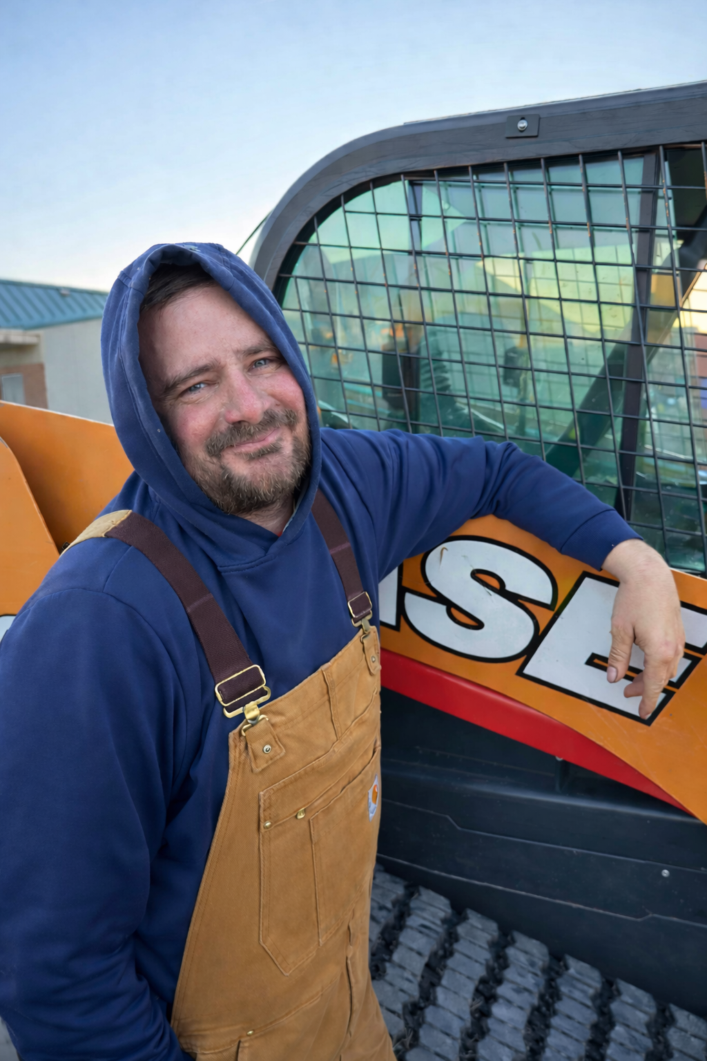 Chad Wilson, owner, wearing a blue hoodie and tan Carhartt overalls smiling outdoors beside a construction or demolition vehicle with a partially visible sign.