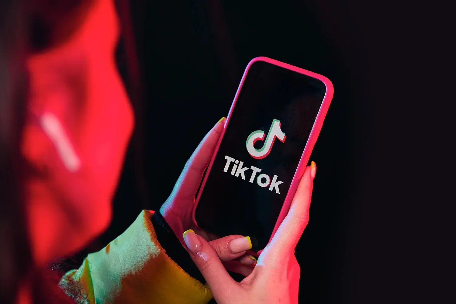 A person holding a smartphone displaying the TikTok app logo on a black background with red and green lighting.