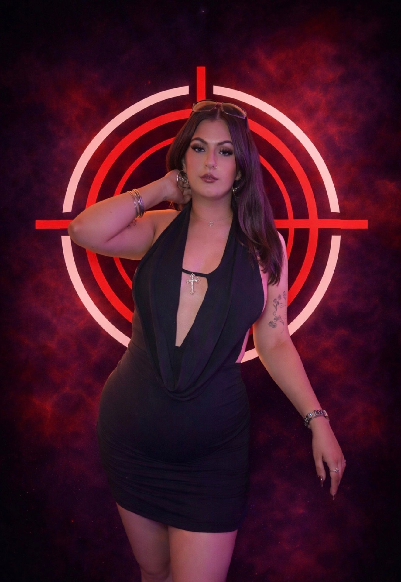 A woman with long dark hair standing in front of a neon red crosshair background, wearing a black dress with a deep neckline and sunglasses on her head.