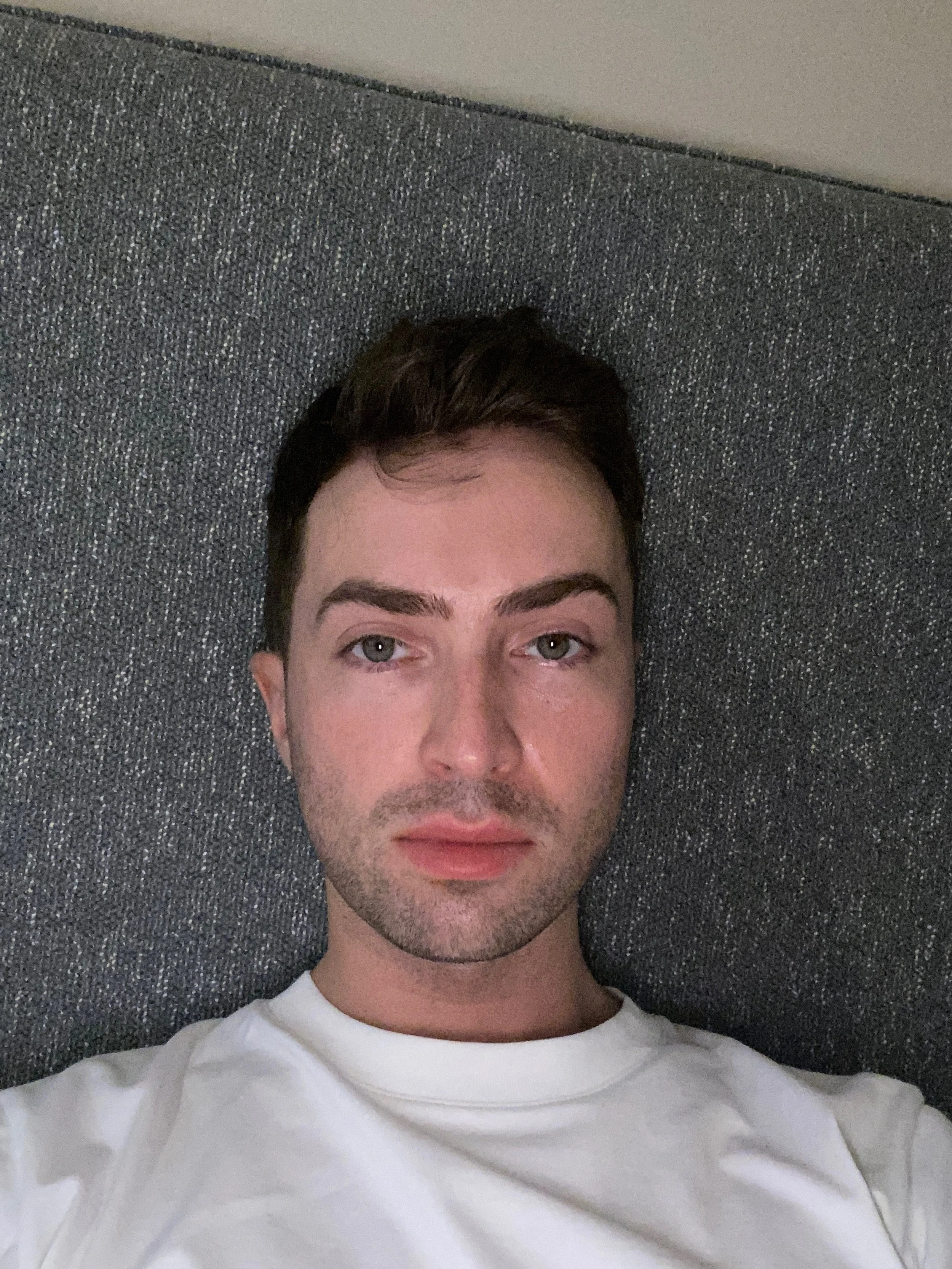 A young man with short dark hair, light skin, and light-colored eyes, lying on a textured gray headboard, wearing a white t-shirt.