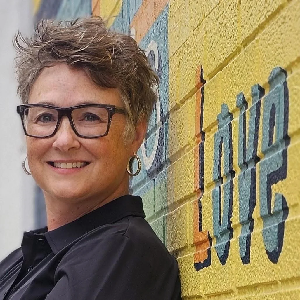 A smiling woman with short curly hair, glasses, and hoop earrings standing next to a colorful mural reading 'Creating Hope' on a brick wall.