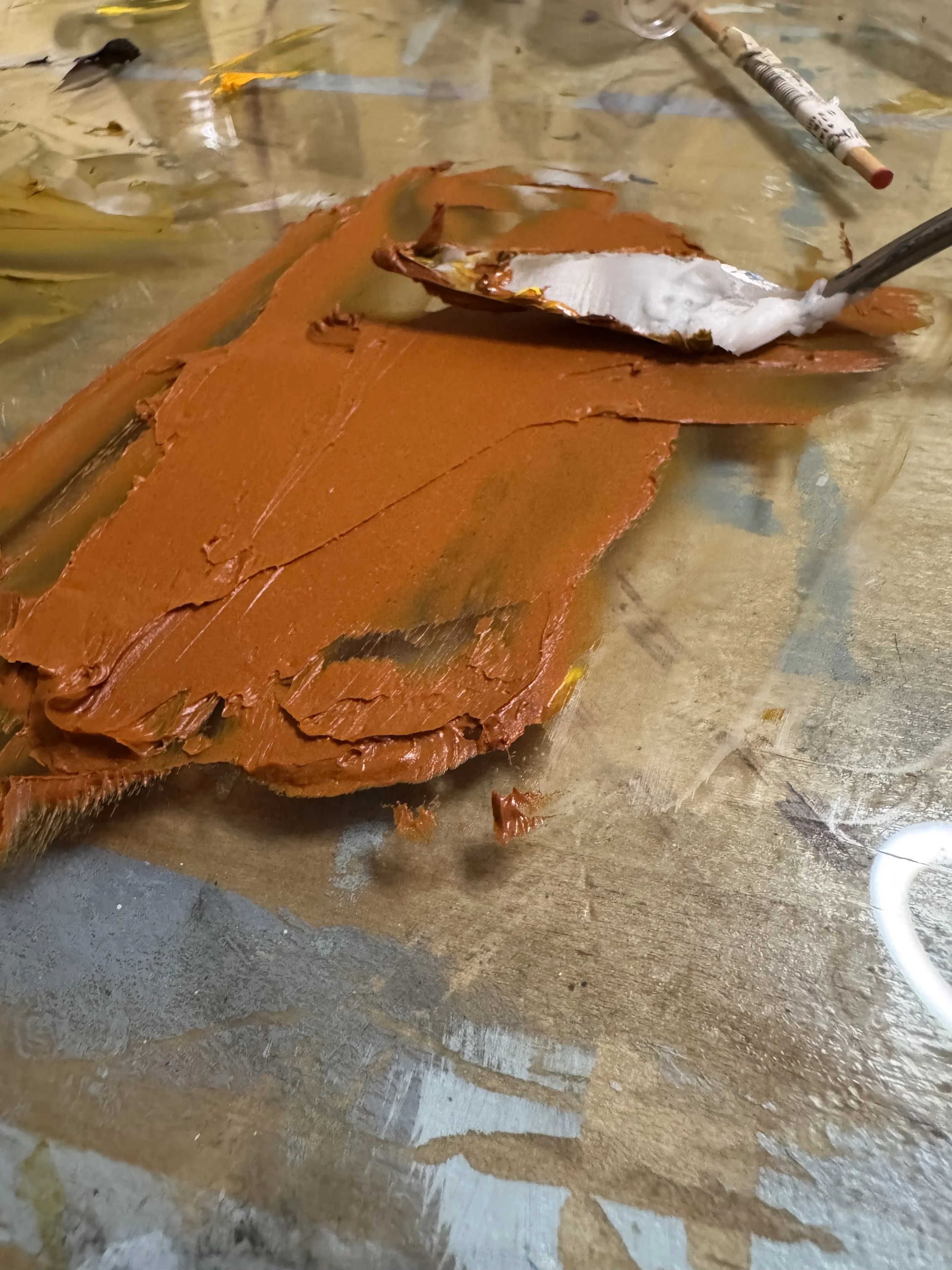 A burnt orange sheet of painted metal or paper on a wooden table with a silver spoon and a paintbrush nearby.