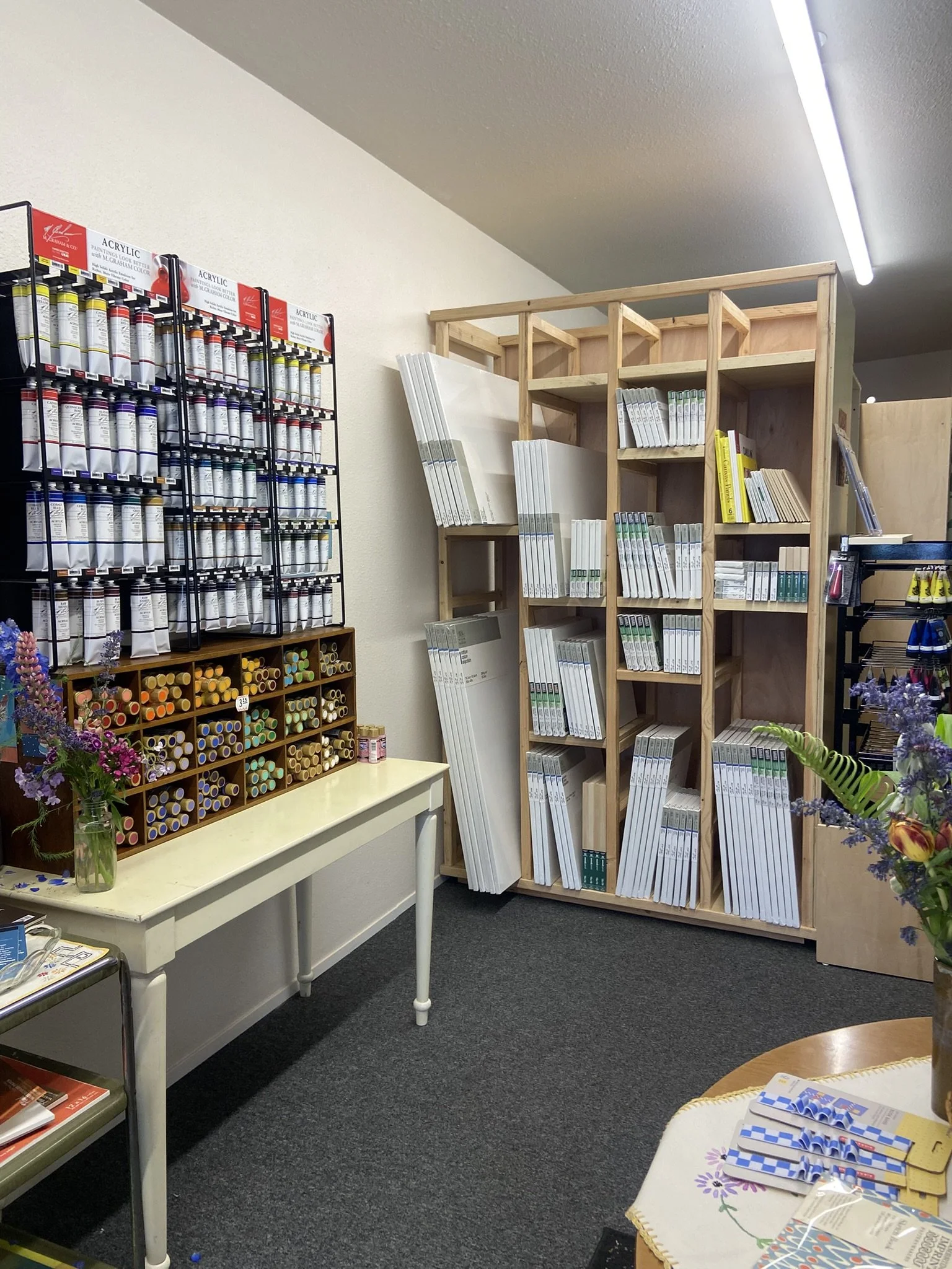 Interior of an office supply store with shelves of books and art supplies, a white table with a flower vase, and a small table with brochures and magazines.
