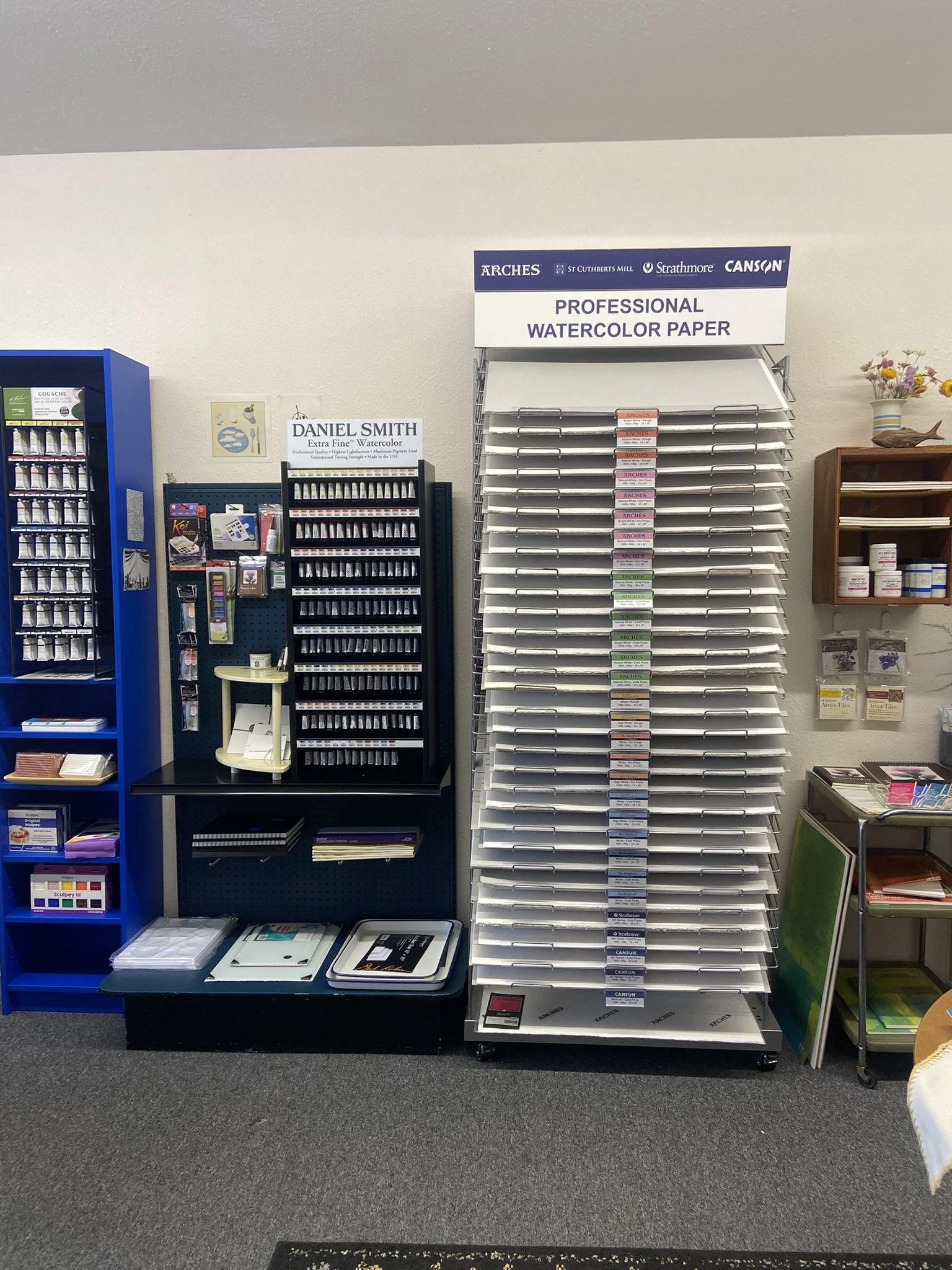 Display of watercolor paper and art supplies in an art store, including a stand with watercolor paper, small shelves with art notebooks and folders, and a vending machine with art products.