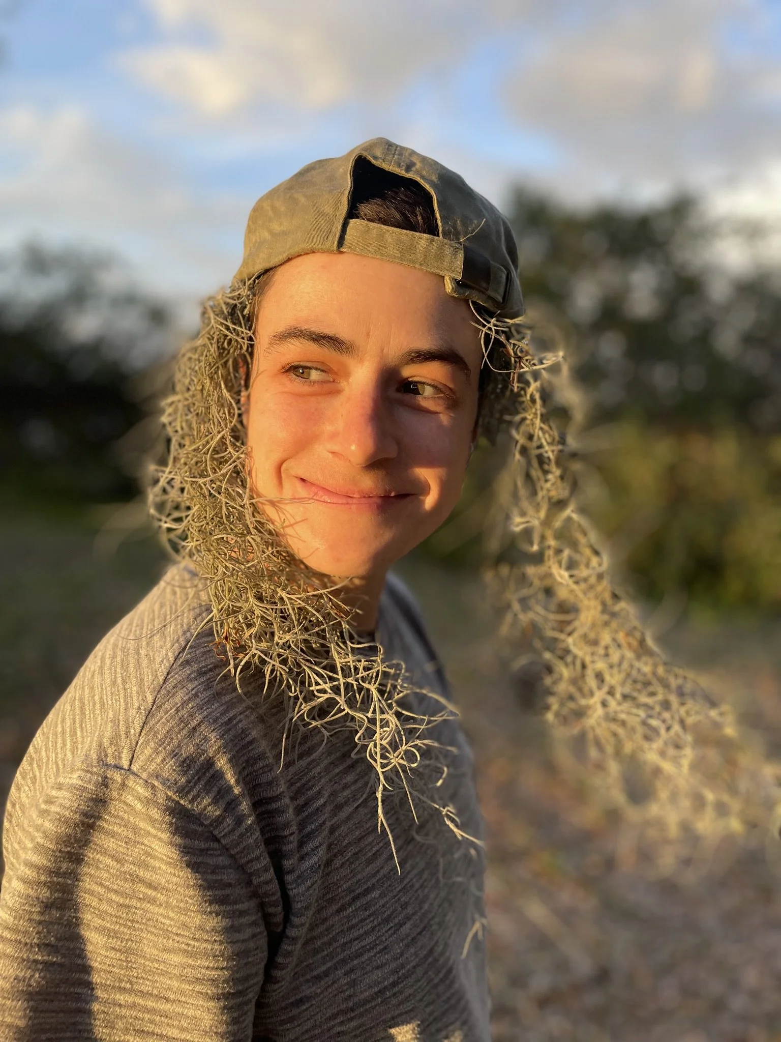 A person smiling outdoors in late afternoon sunlight, wearing a gray sweatshirt and a backwards baseball cap, with dry branches around their head.