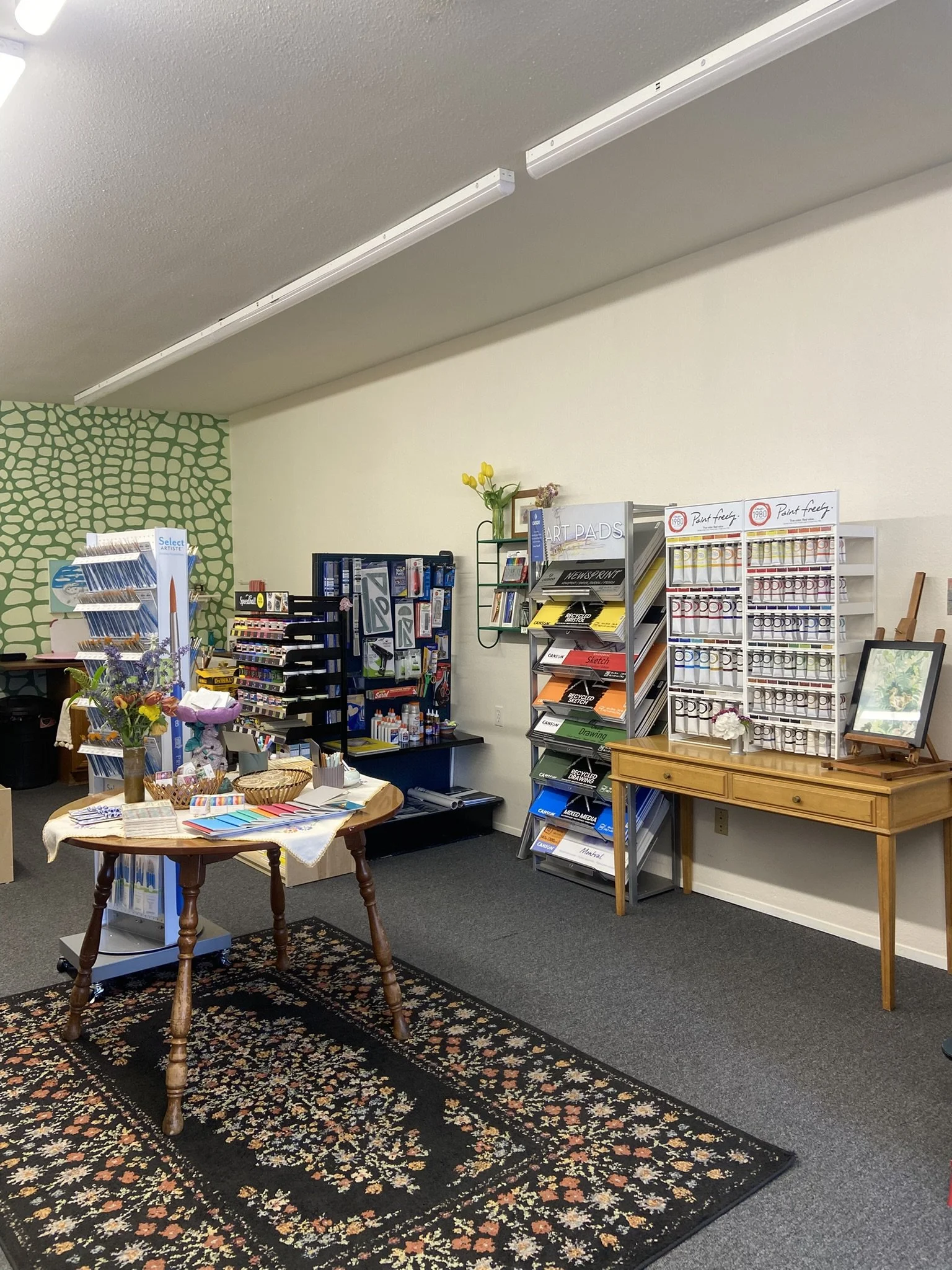 Interior of an office or store with shelves of art supplies and office materials, a table with presents and flowers, and a floral rug on the carpeted floor.