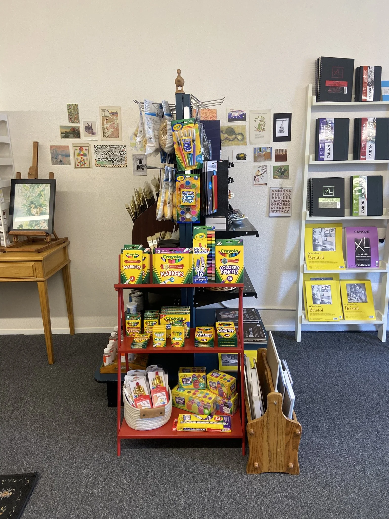 A display of art supplies, including markers, colored pencils, paint sticks, and craft items, arranged on a black and red shelving unit in an art store. To the right, there is a white shelf with art notebooks and pads, and to the left, a small wooden side table and framed artwork leaning against the wall. The wall behind has various small art prints and illustrations attached.