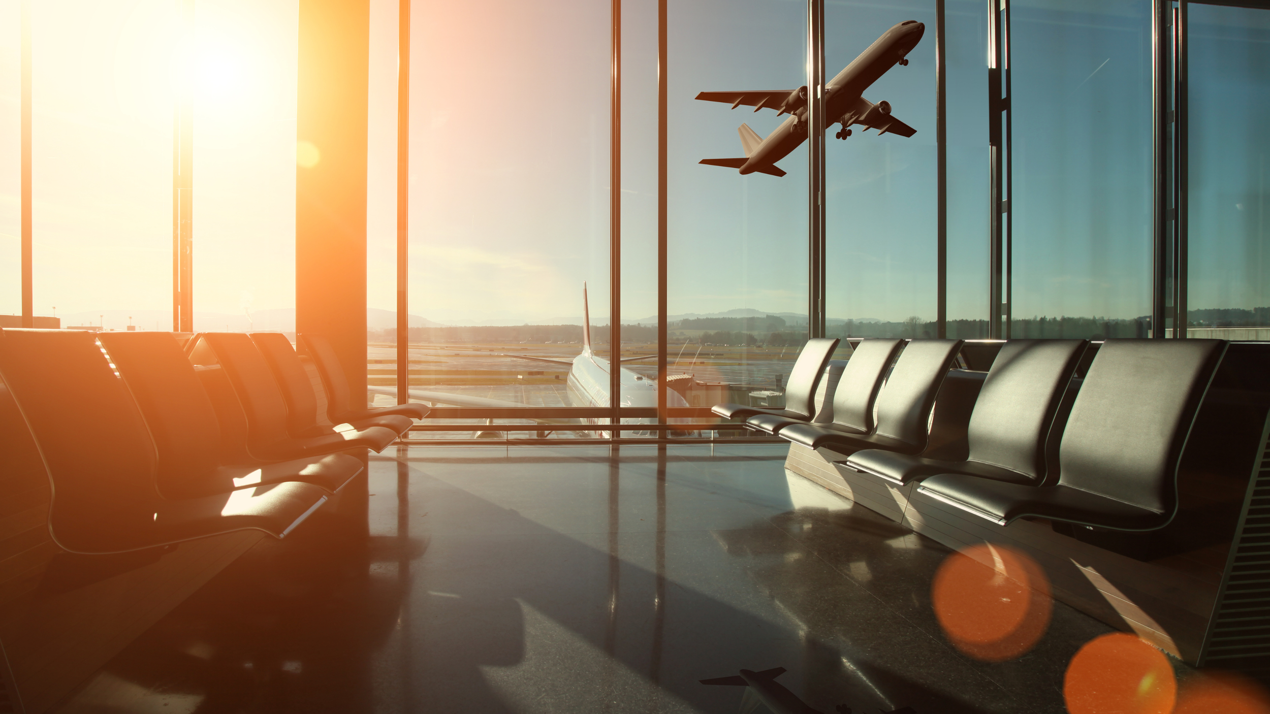 Airport waiting area with empty black chairs, large glass windows showing a plane taxiing outside, and an airplane flying overhead during sunrise or sunset.