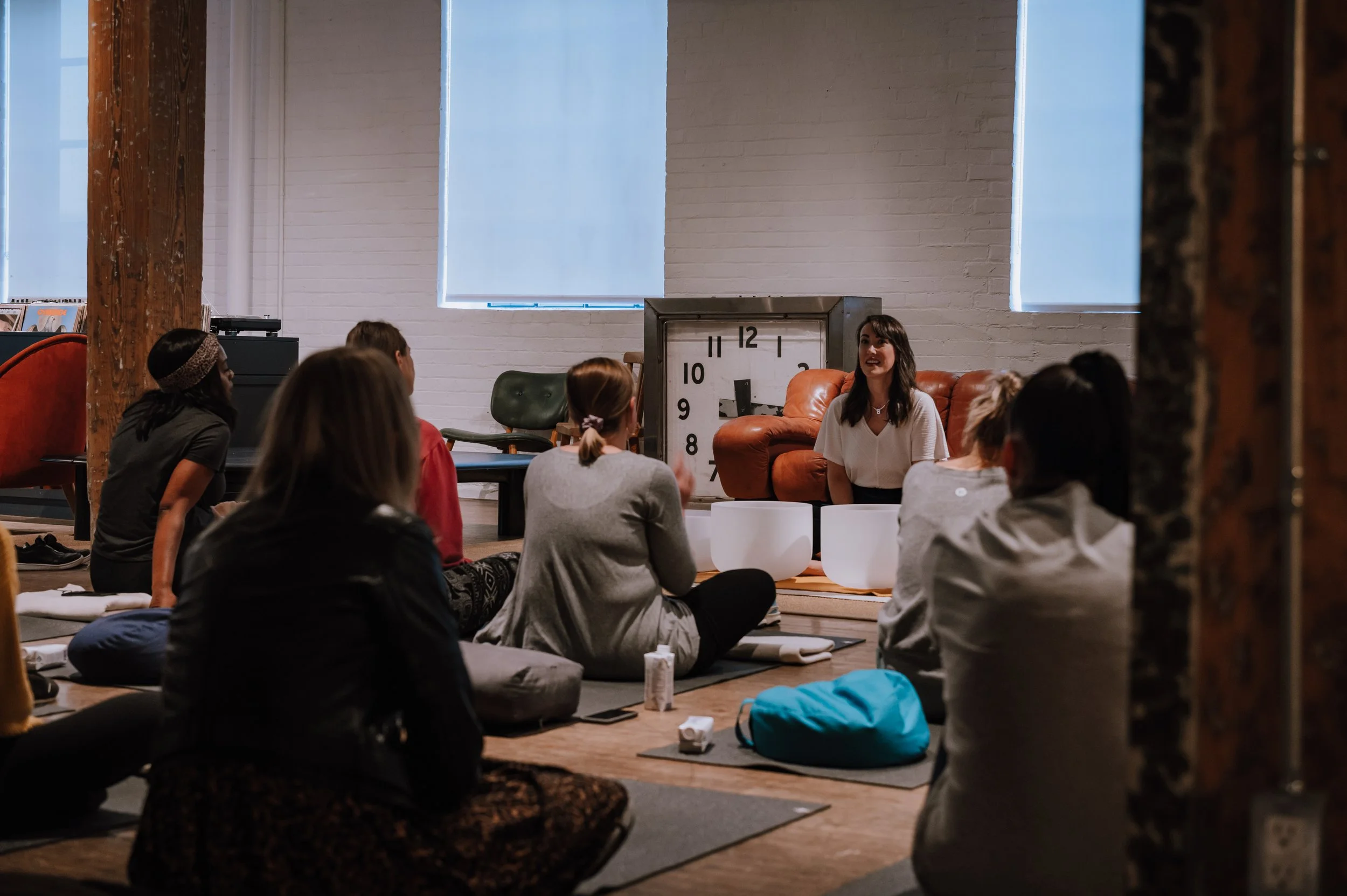 A group of women participating in a circle meditation or discussion session in a cozy, industrial-style room with white brick walls and large windows, centered around a woman leading the session, seated near a large clock and an orange couch.