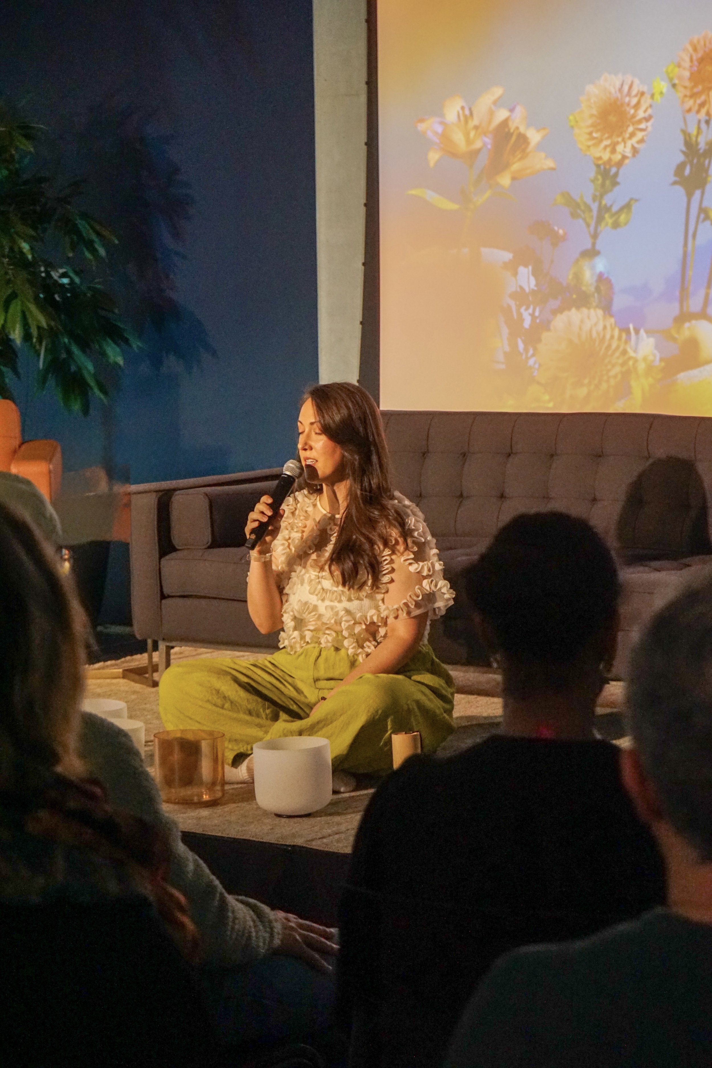 A woman is sitting on the floor with a microphone, her eyes closed, speaking or singing during an event. Behind her is a large screen displaying a picture of flowers in vases. There are audience members in front of her, and a cozy setting with couches, plants, and ambient lighting.