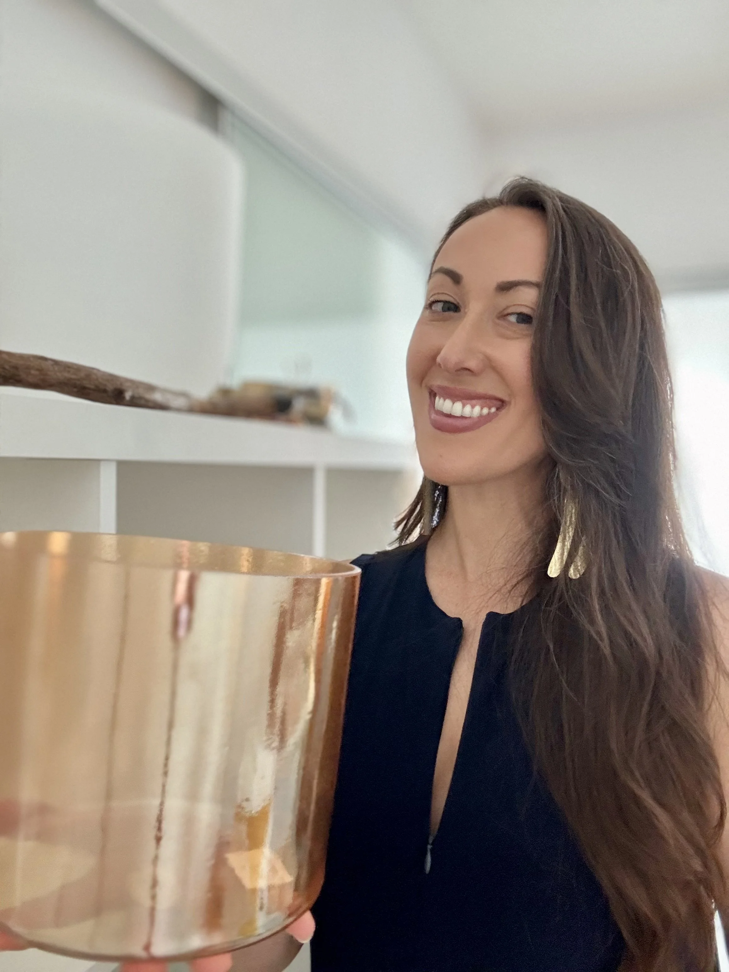 A woman with long brown hair, smiling, holding a gold-colored candle holder, indoors with light-colored walls and furniture.