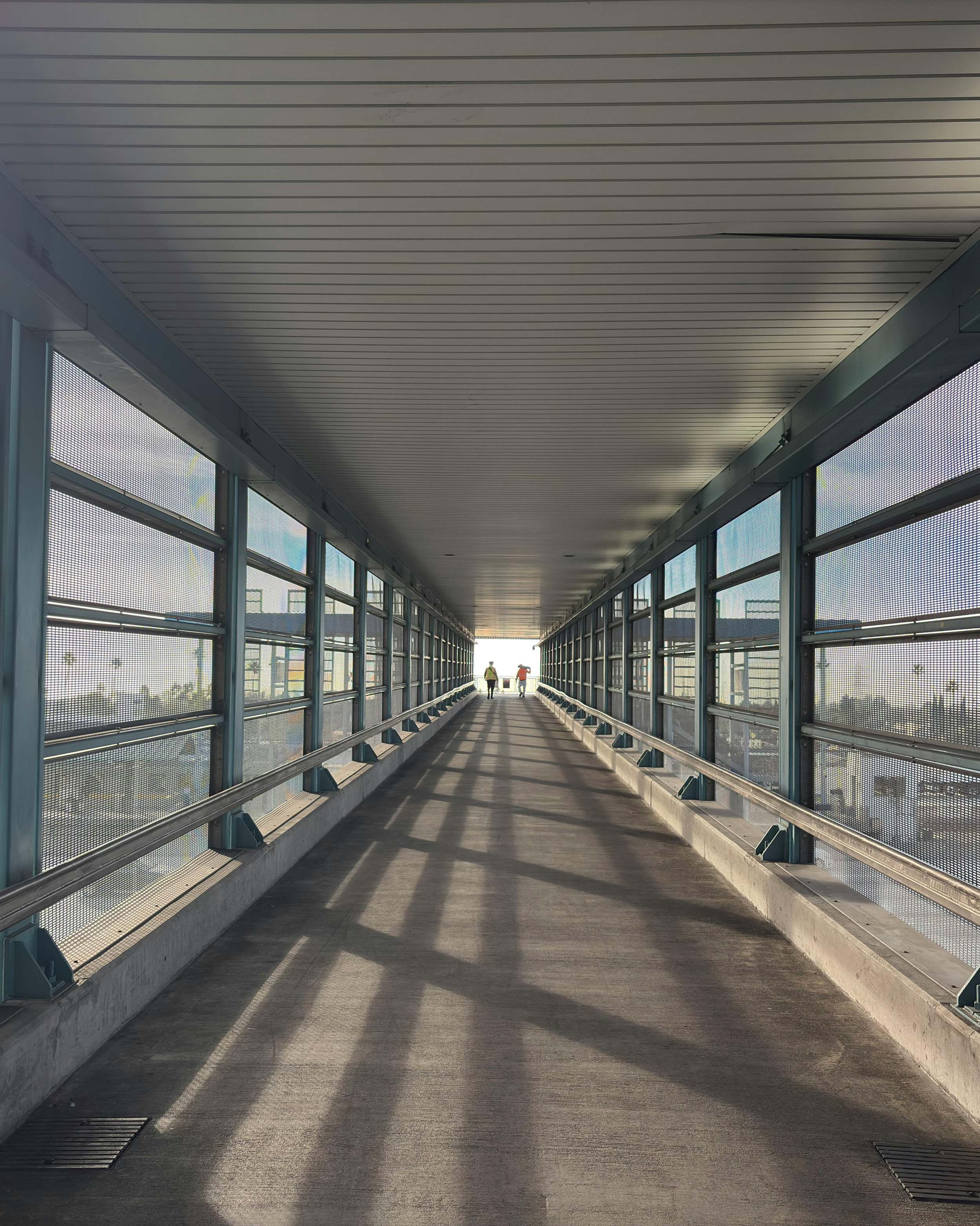 Elevated walkway at an airport or transit station with glass walls, shadows on the floor, and two people walking towards the exit