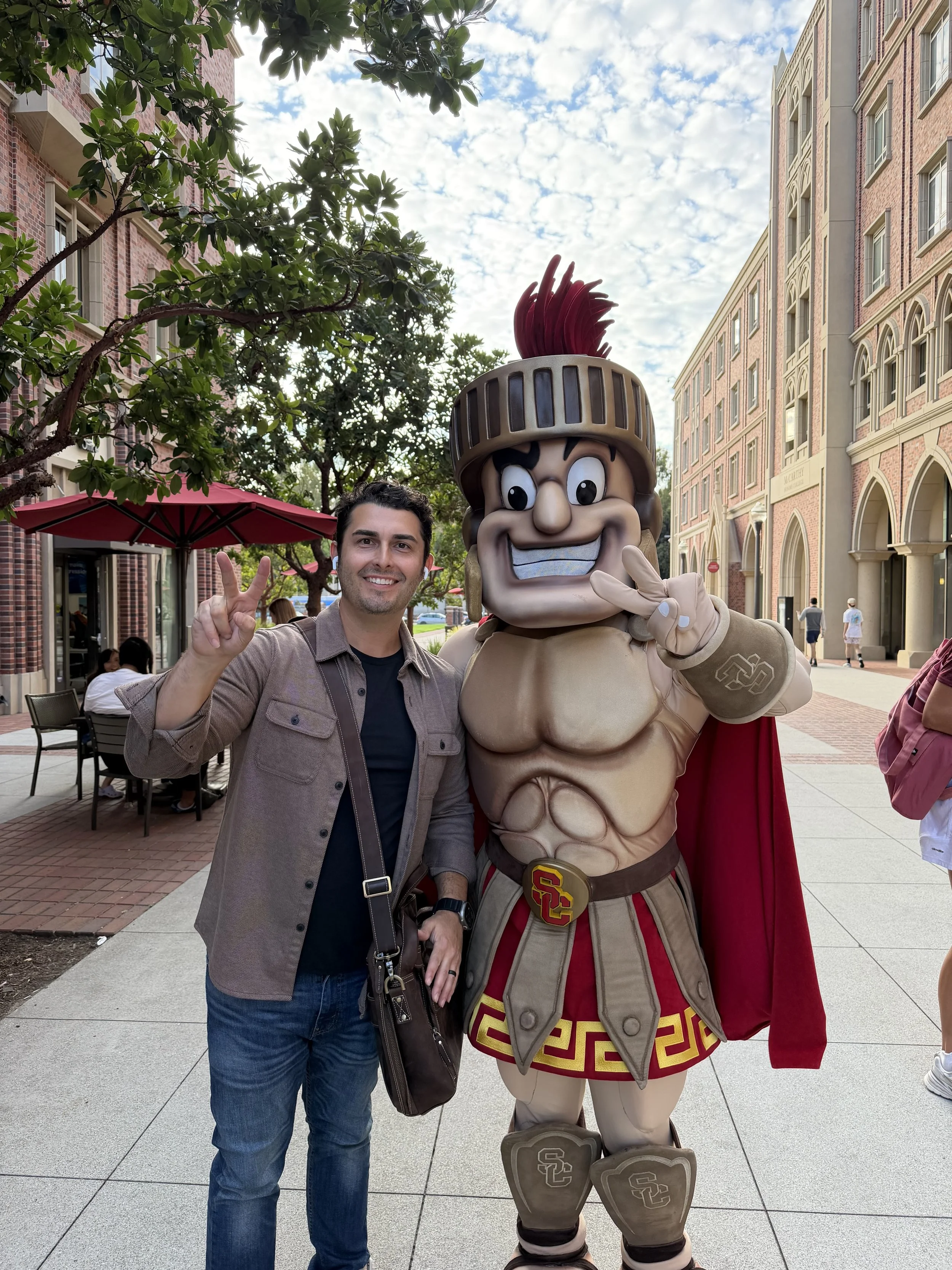 A man smiling and making a peace sign pose next to a person dressed as a cartoon warrior character with a large helmet, a big smile, and a red cape, standing outdoors in a city walk area with brick buildings and trees.