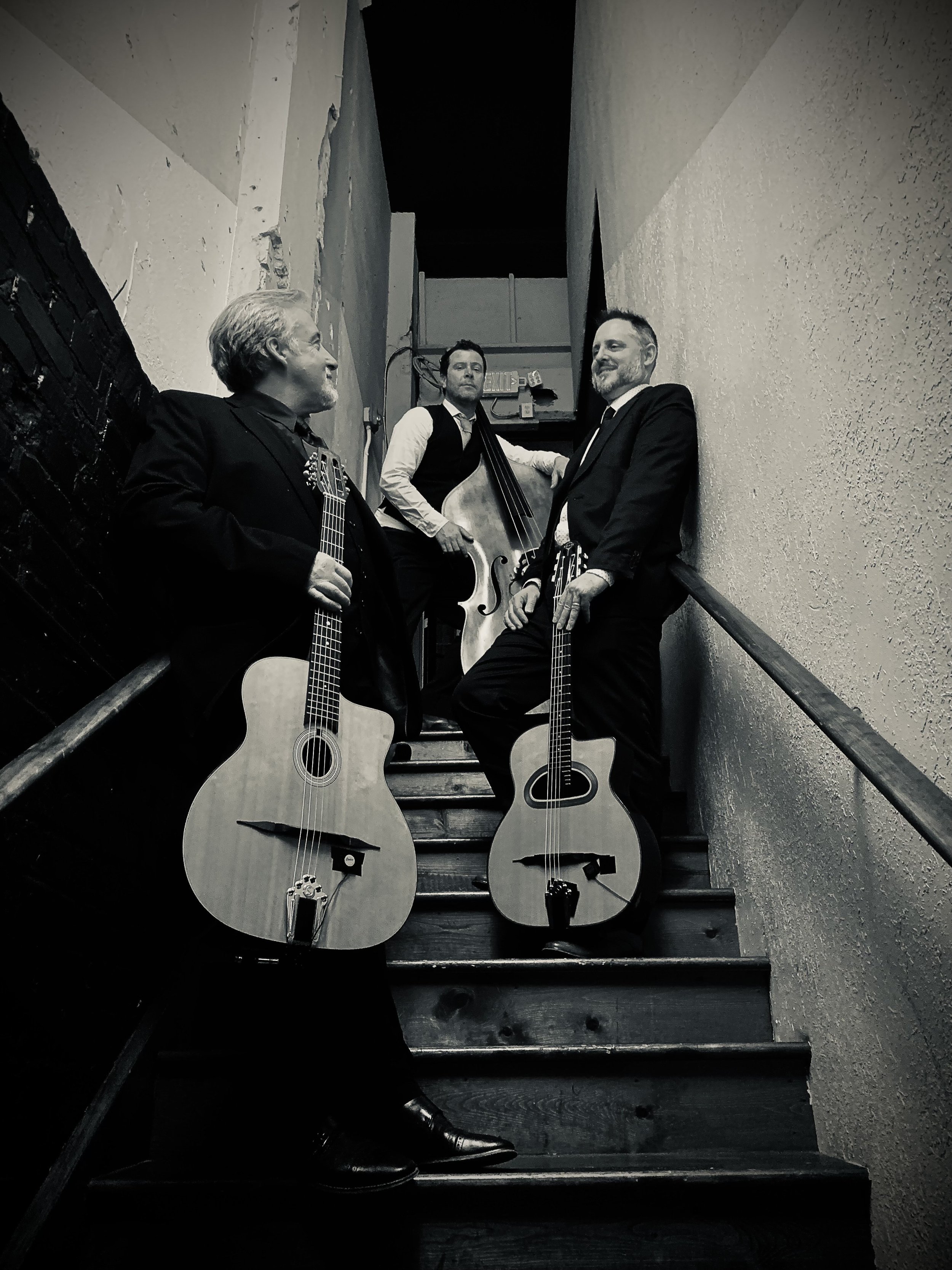 Black and white photo of three men in suits holding guitars, standing on a staircase in a narrow corridor.