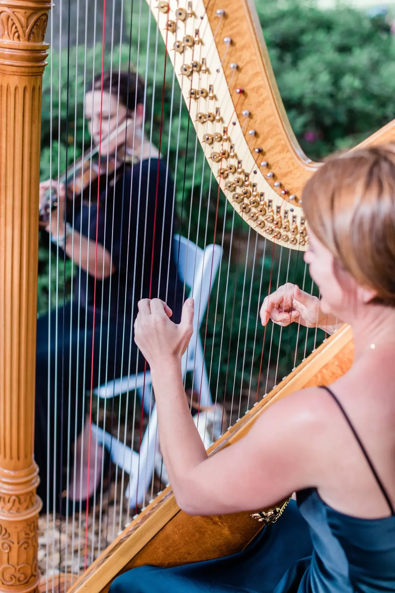 A woman playing a golden harp outdoors, with another woman sitting on a white chair in the background, blurred, and green foliage around them.